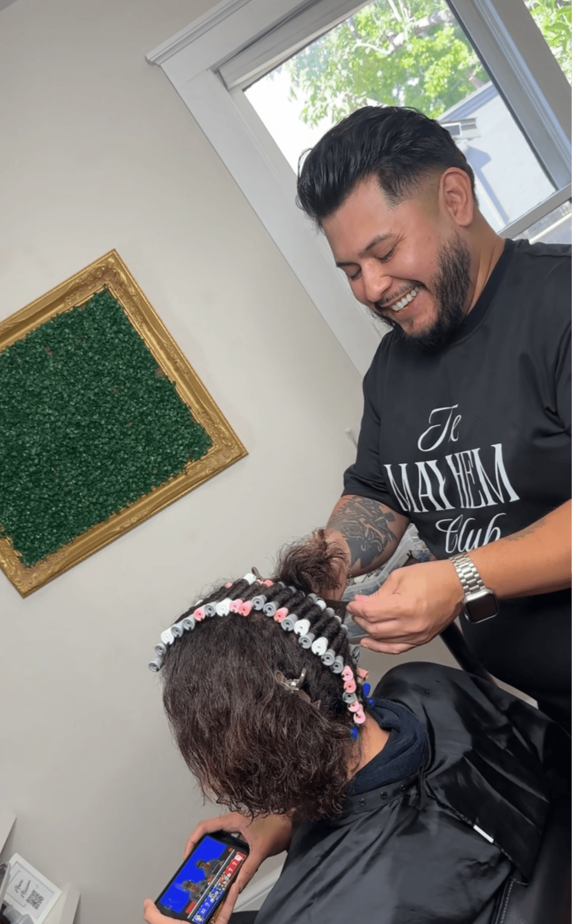 Man with a beard and tattooed arm smiling while styling a child's hair with beads, in a room with a window and green wall art.