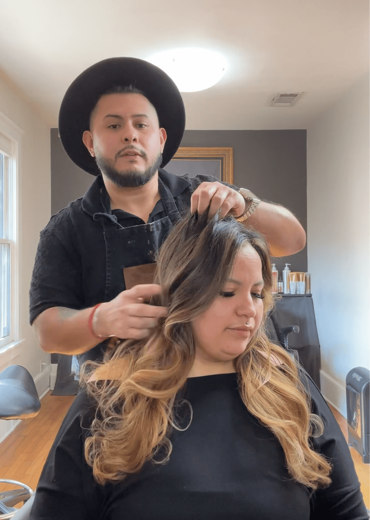 A hairstylist with a black hat and black shirt styles a woman's long, wavy hair in a room with gray walls.