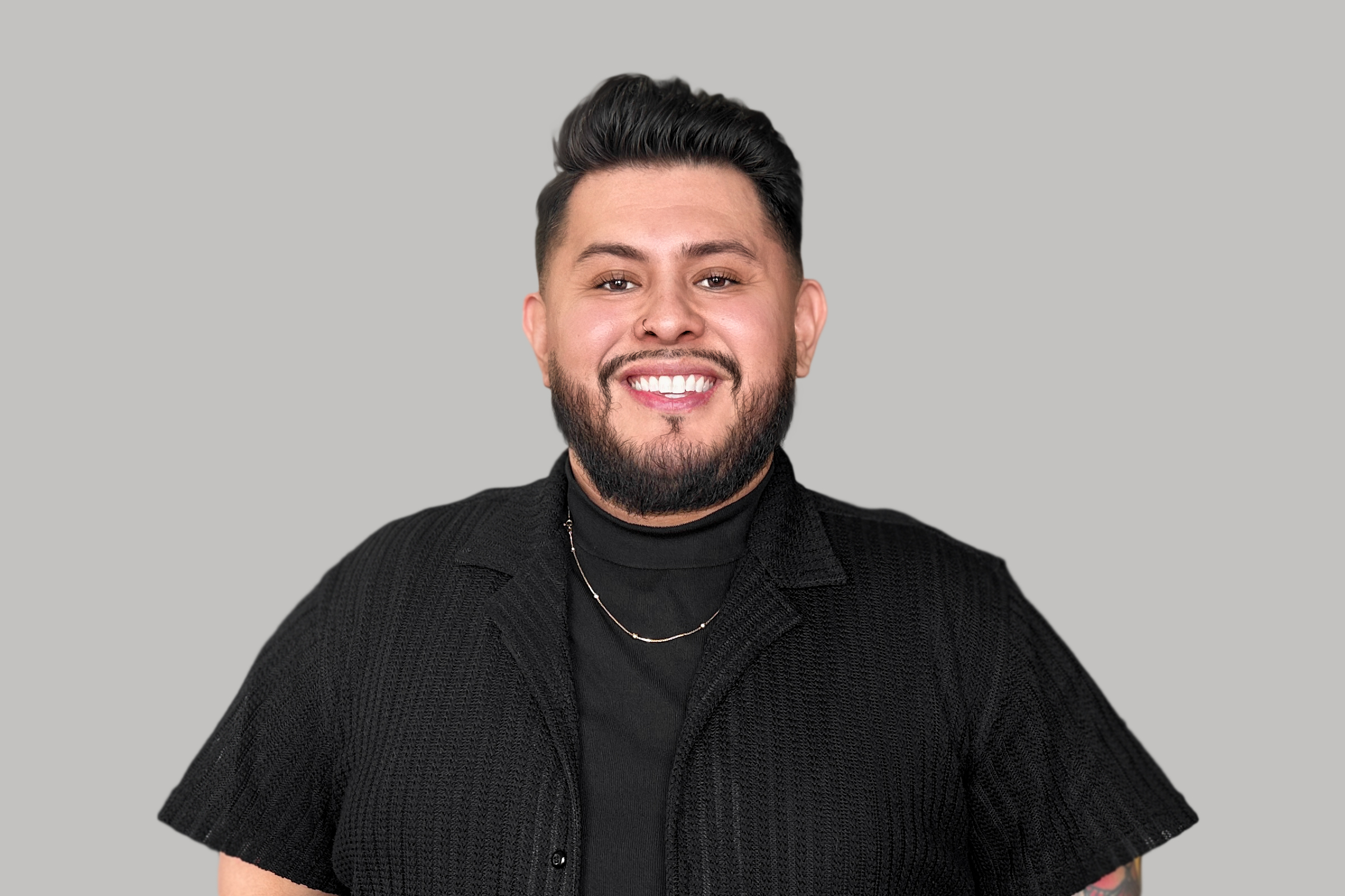 Smiling man with dark hair and beard wearing black shirt and necklace, gray background.
