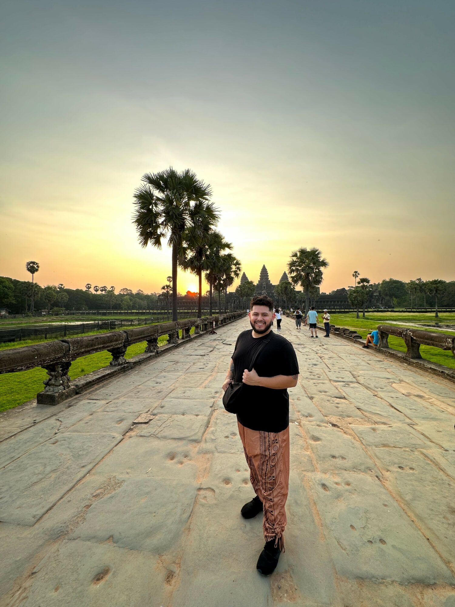 Person standing on a paved path with trees and a sunset sky in the background.