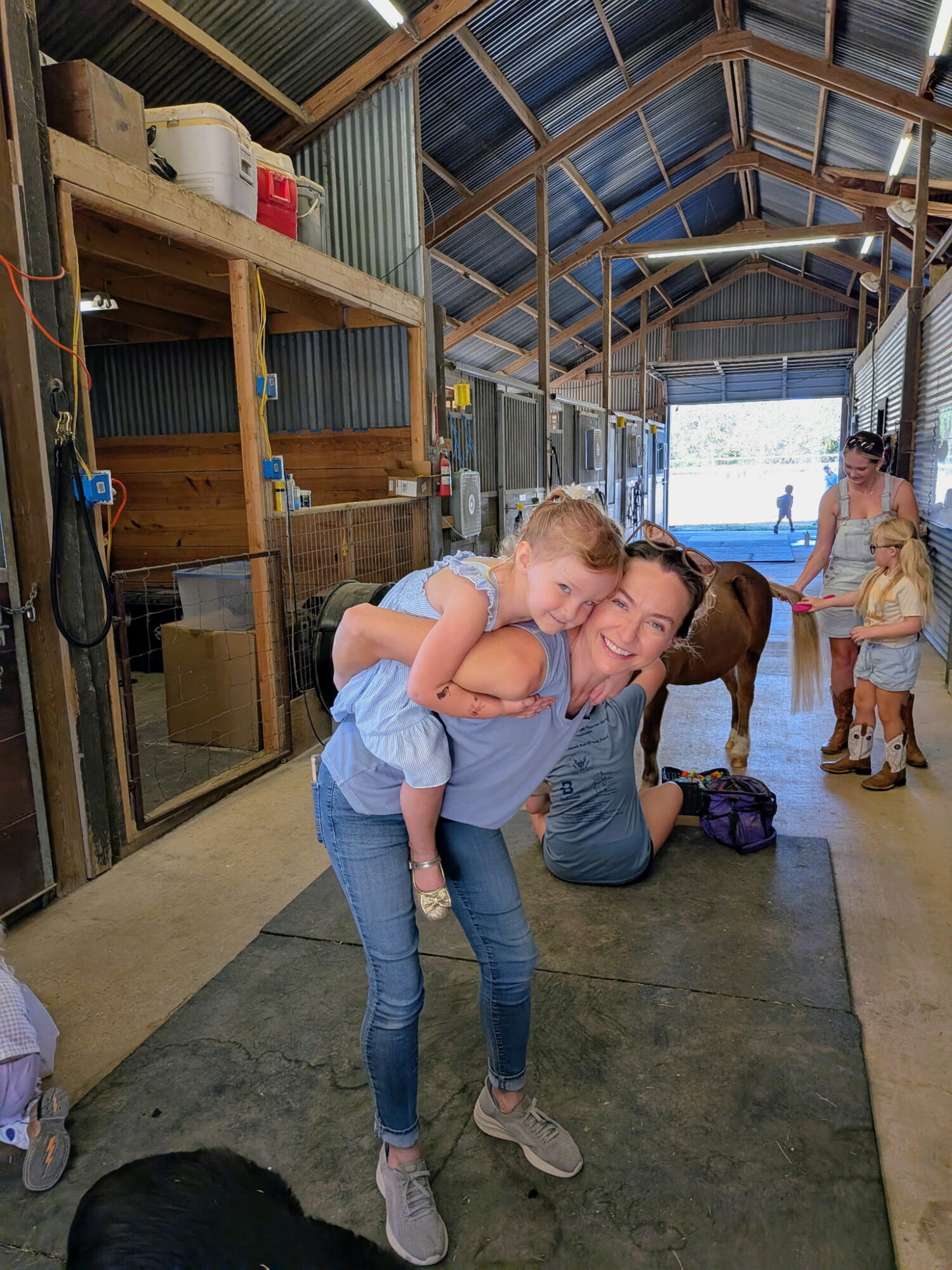 Child being carried on an adult's back inside a barn with people and animals, wooden shelves, and a high arched ceiling.
