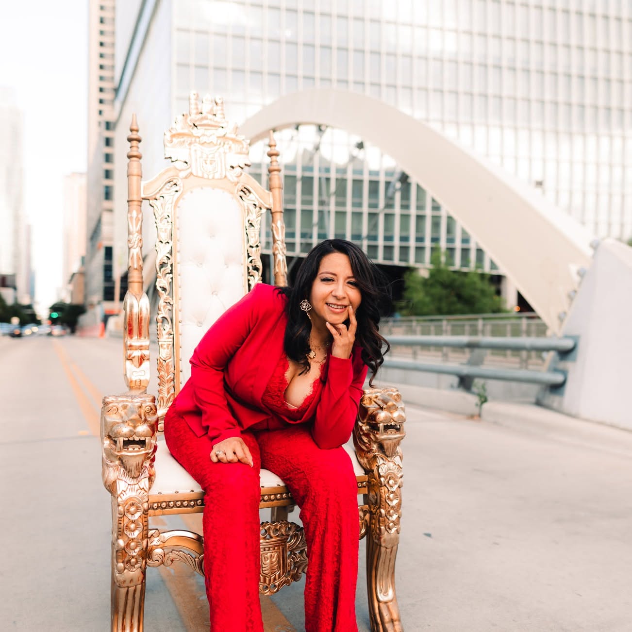 Woman in red outfit sitting on ornate throne-like chair outdoors, smiling, city buildings in background.