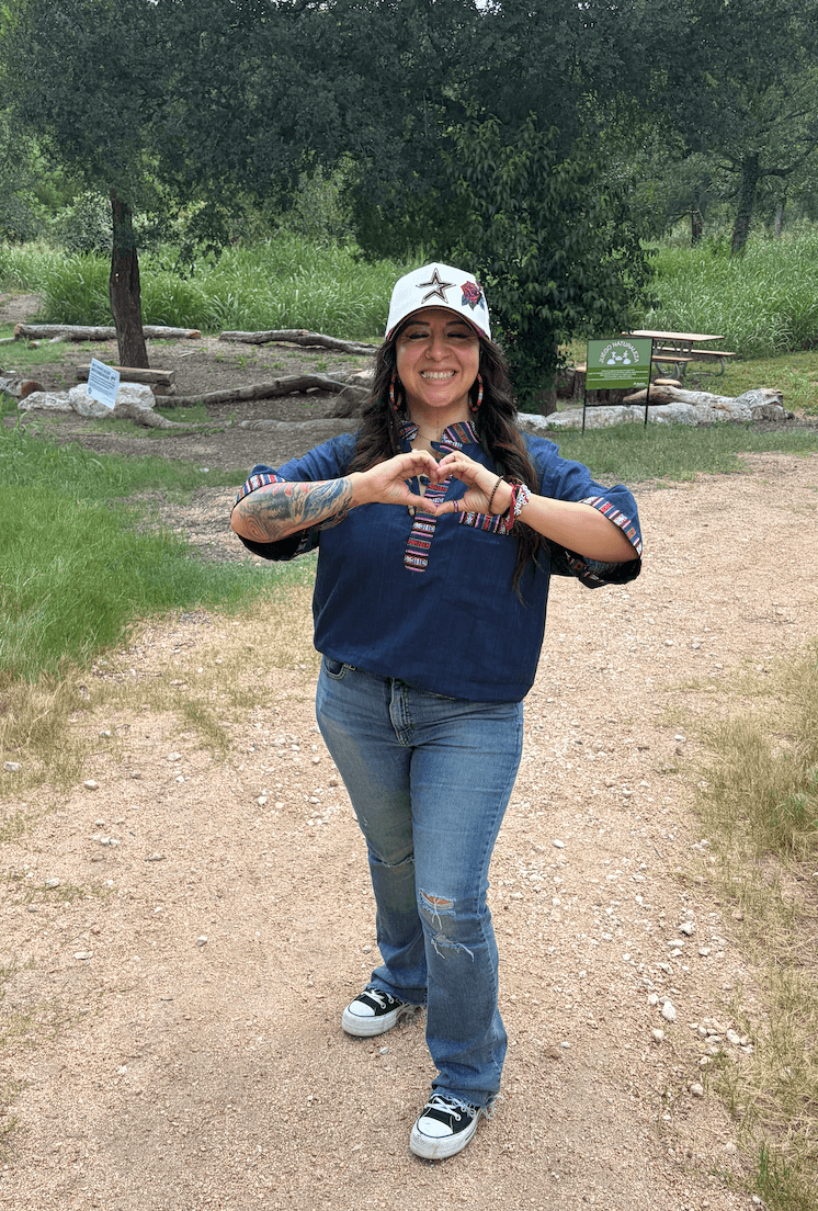 Woman standing outdoors on a dirt path, smiling, wearing a baseball cap, blue shirt, jeans, and sneakers, making a heart shape with her hands.