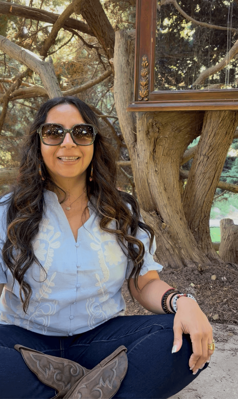 Woman with long dark hair wearing sunglasses, a light blue shirt, and dark pants, sitting outdoors near a tree and a window frame.