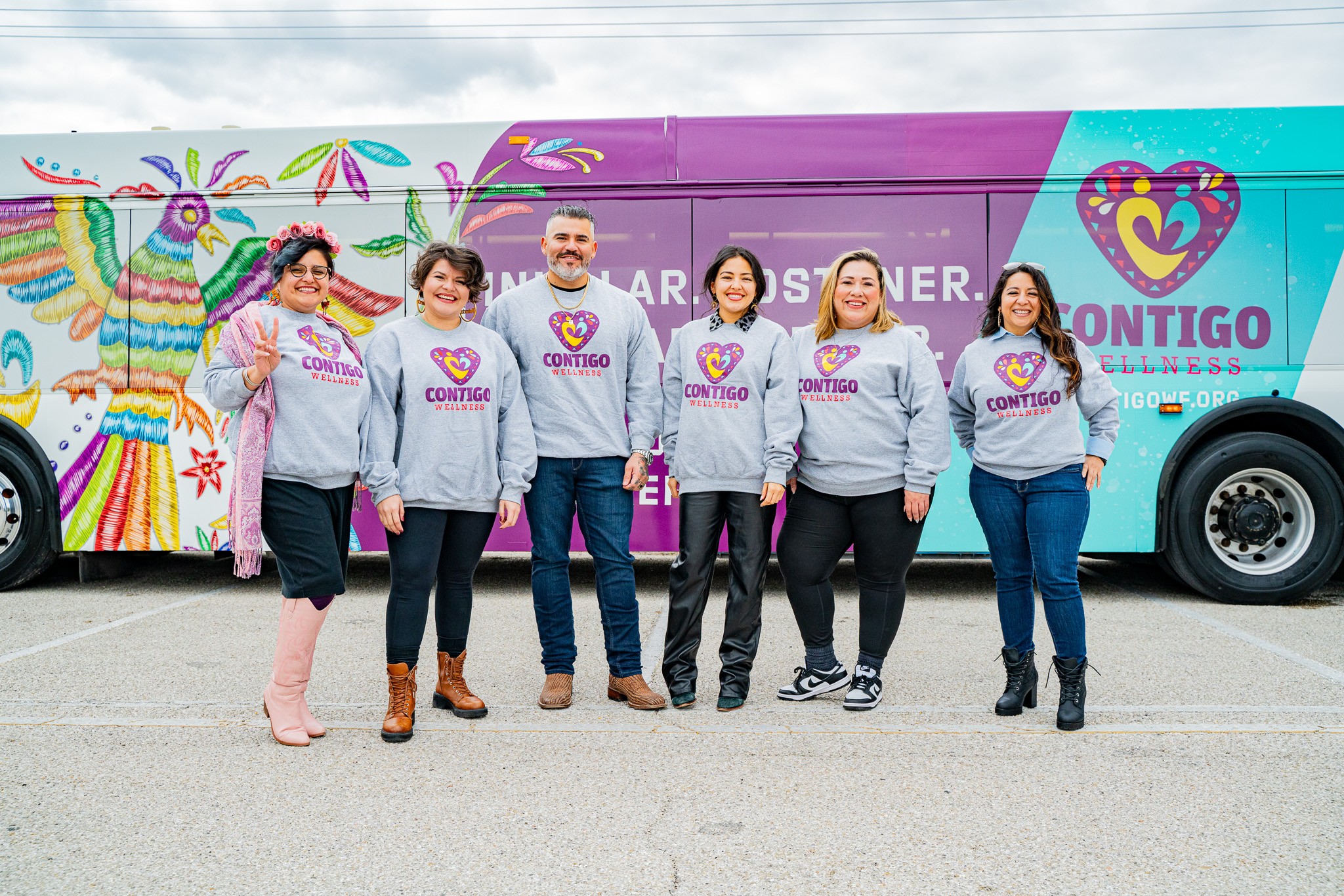 Six people stand in front of a colorful bus with a heart logo and the word 'Contigo' on it, smiling.