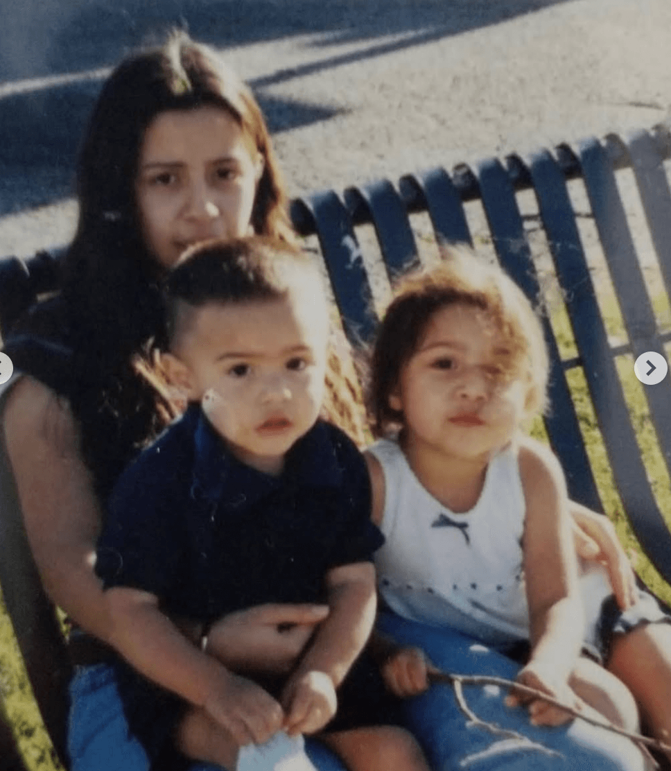 Three children sitting outdoors on a bench, with a fence and sunlight in the background.
