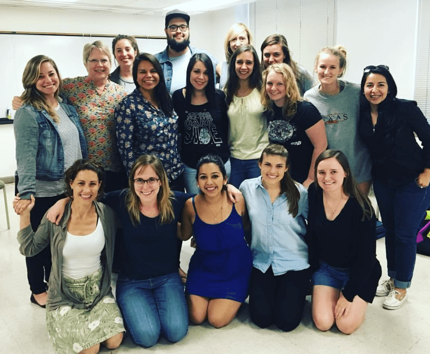 Group of 16 people, mostly women, standing and sitting indoors, smiling, in casual clothing, with a white wall background.