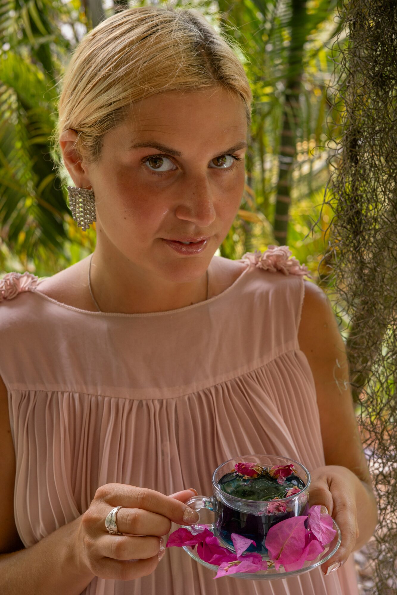 Woman holding a glass dish with dark dessert and pink flower petals, outdoors with green foliage background.
