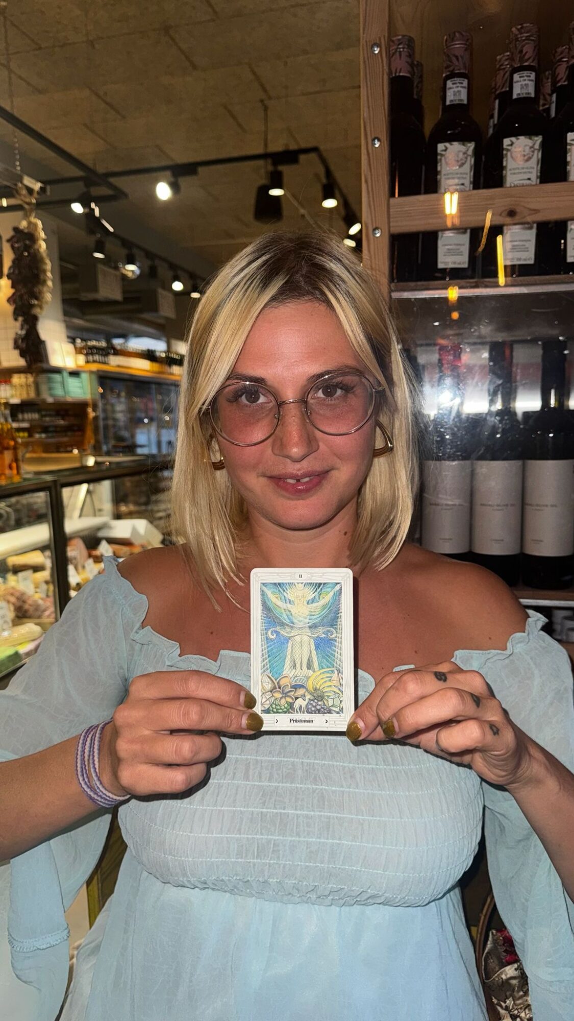 Woman with blonde hair and glasses holding a tarot card in a store or cafe setting.