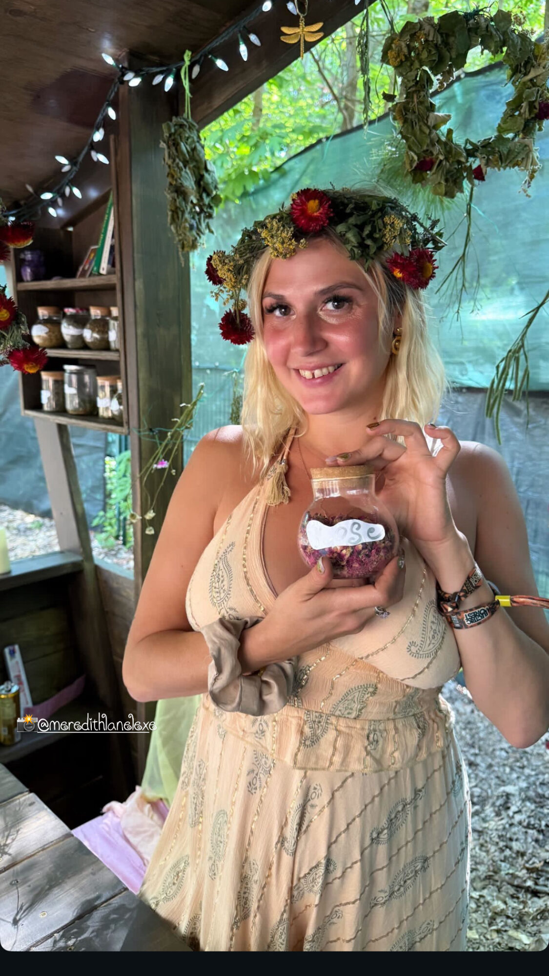 Woman with a flower crown holding a jar, smiling indoors with greenery in the background.