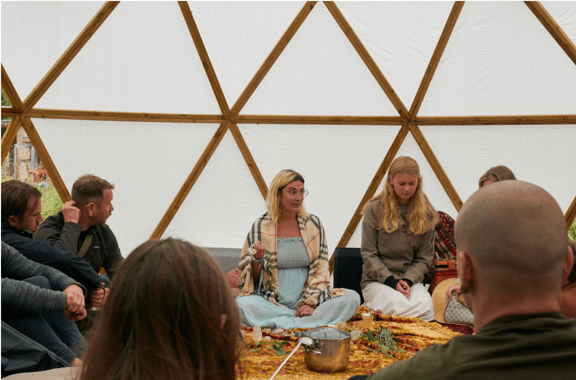 Group of people sitting inside a geodesic dome, listening to a woman speaking, with a pot and food on the floor.