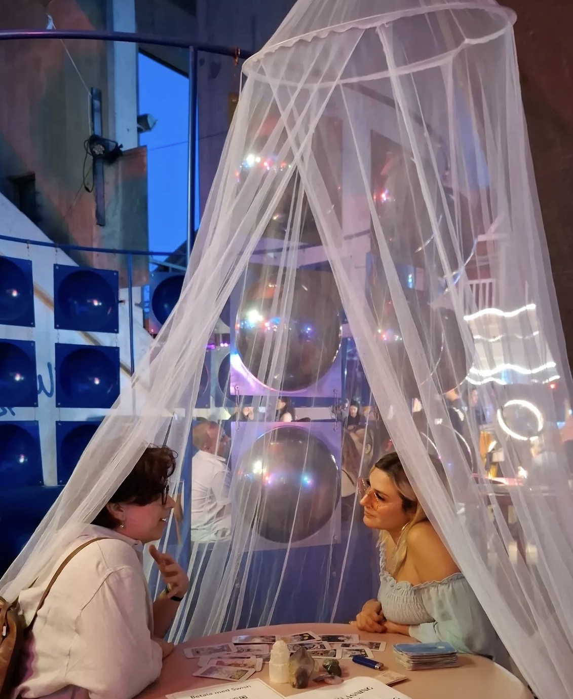Two women sit at a table under a sheer canopy with hanging spheres, engaging in conversation in a colorful setting.