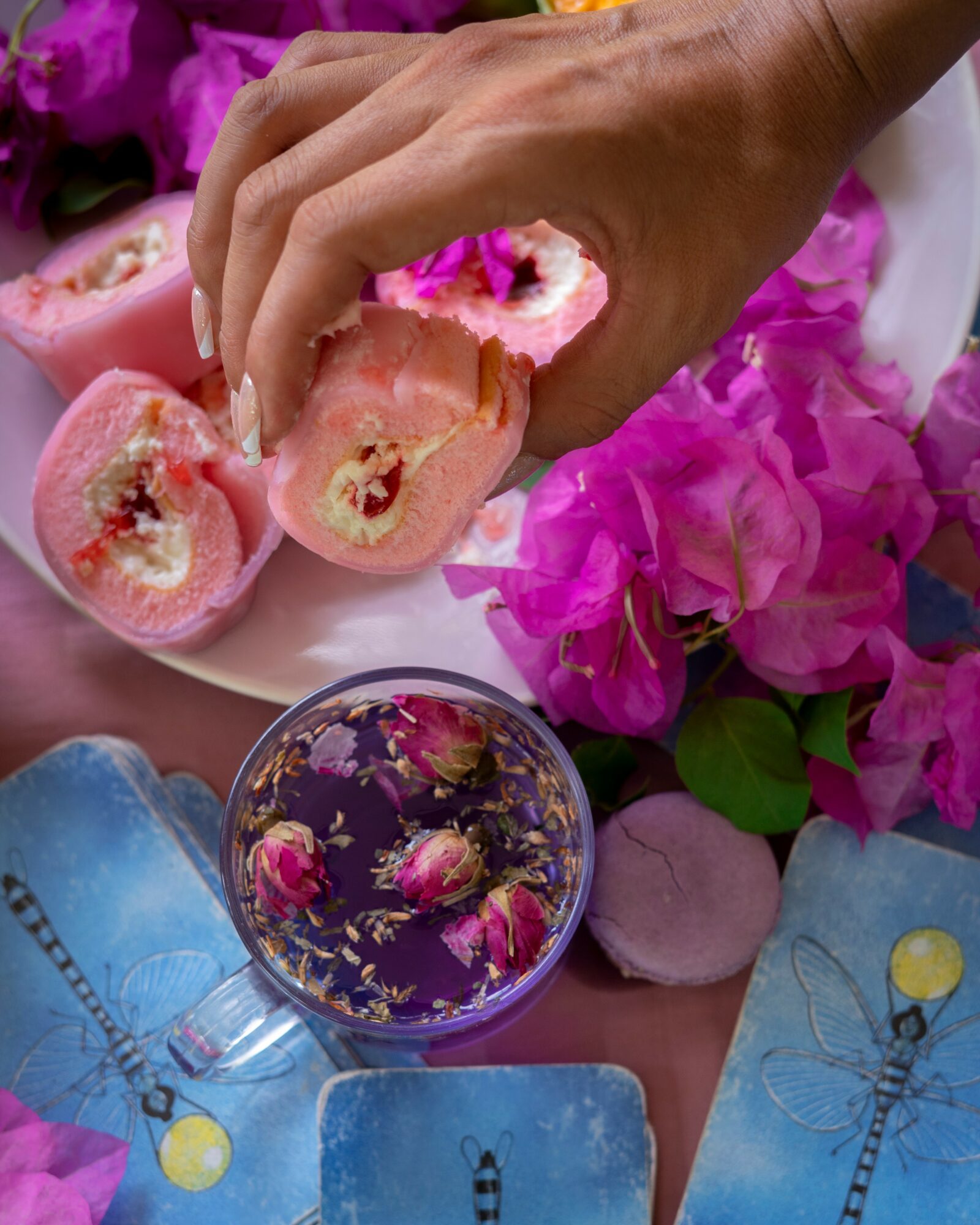 Hand holding a pink dessert with cream and fruit, surrounded by pink flowers and a glass of purple liquid with flowers.