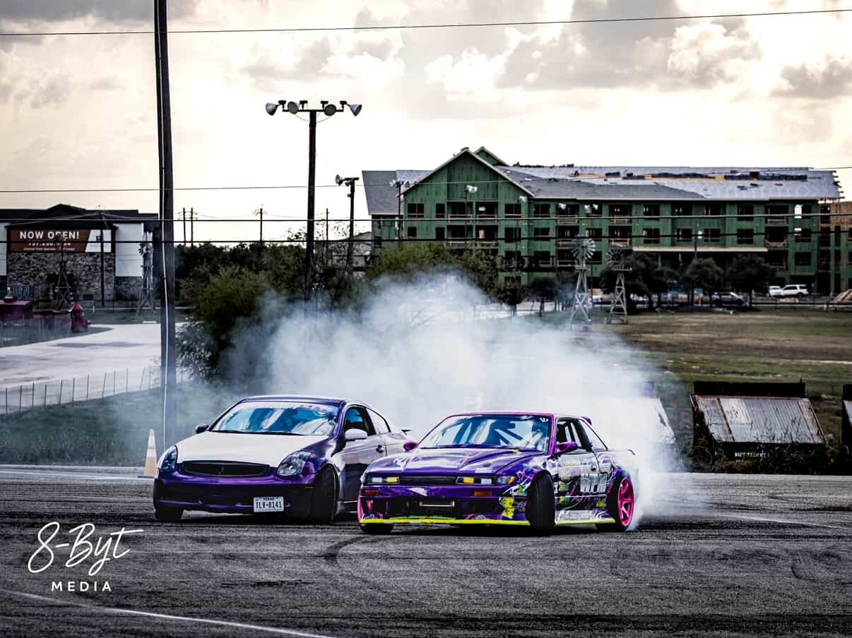 Two race cars drifting on a track with smoke, a building, and cloudy sky in the background.