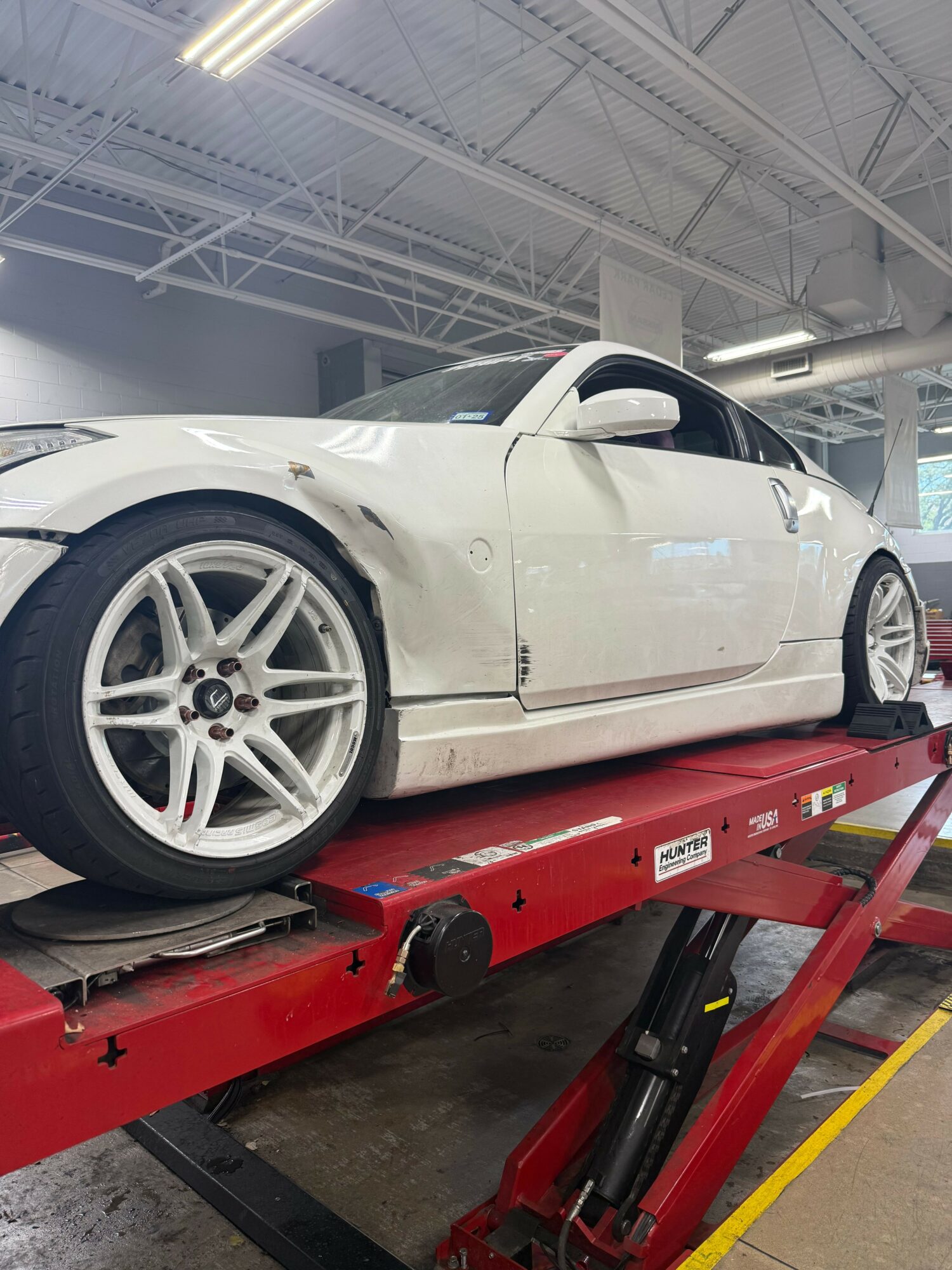 White sports car on a red hydraulic lift in a garage, viewed from the side, with a large wheel and tire visible.