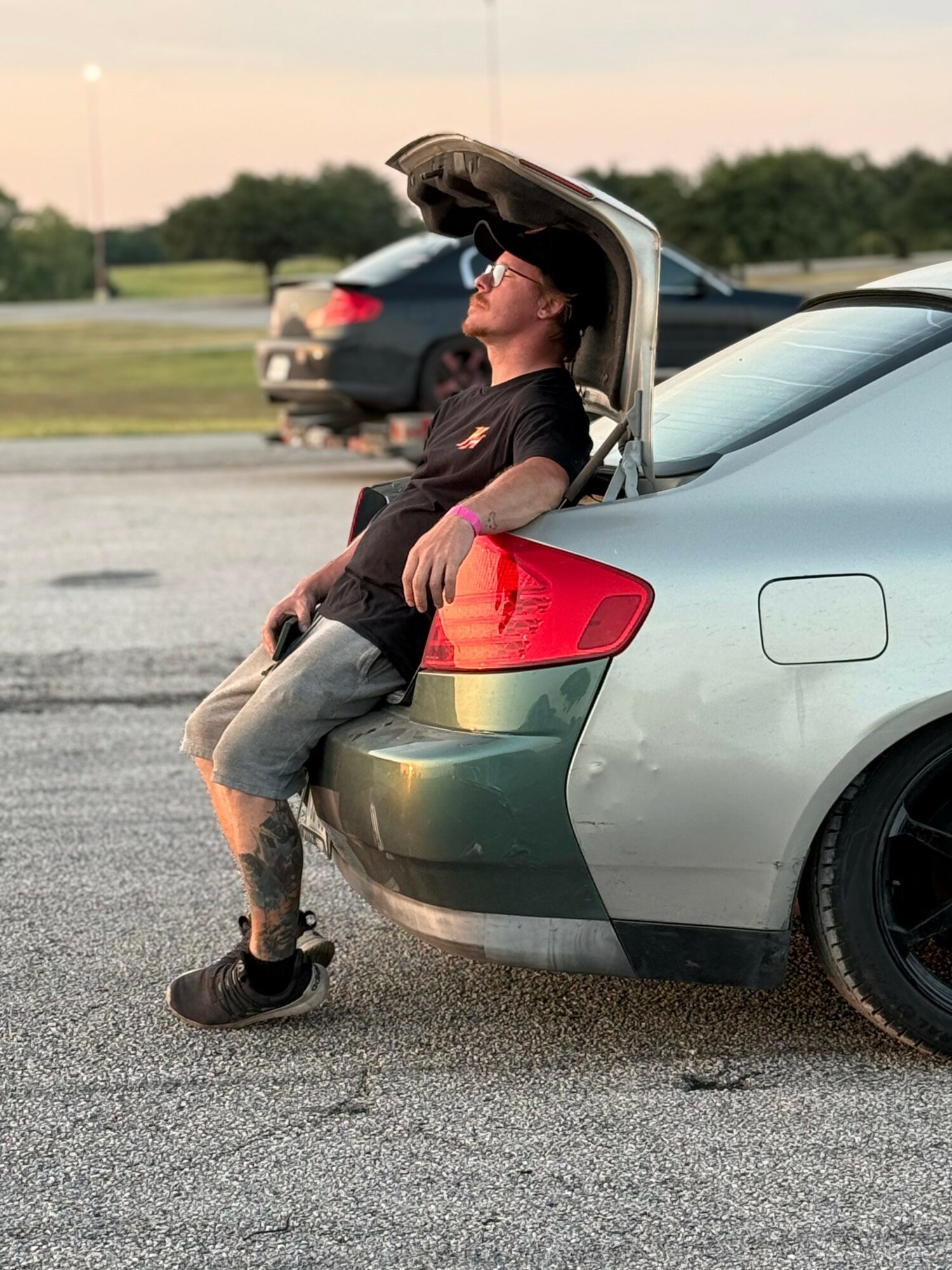 Person leaning against open trunk of a silver car in a parking lot, with other cars and trees in background.