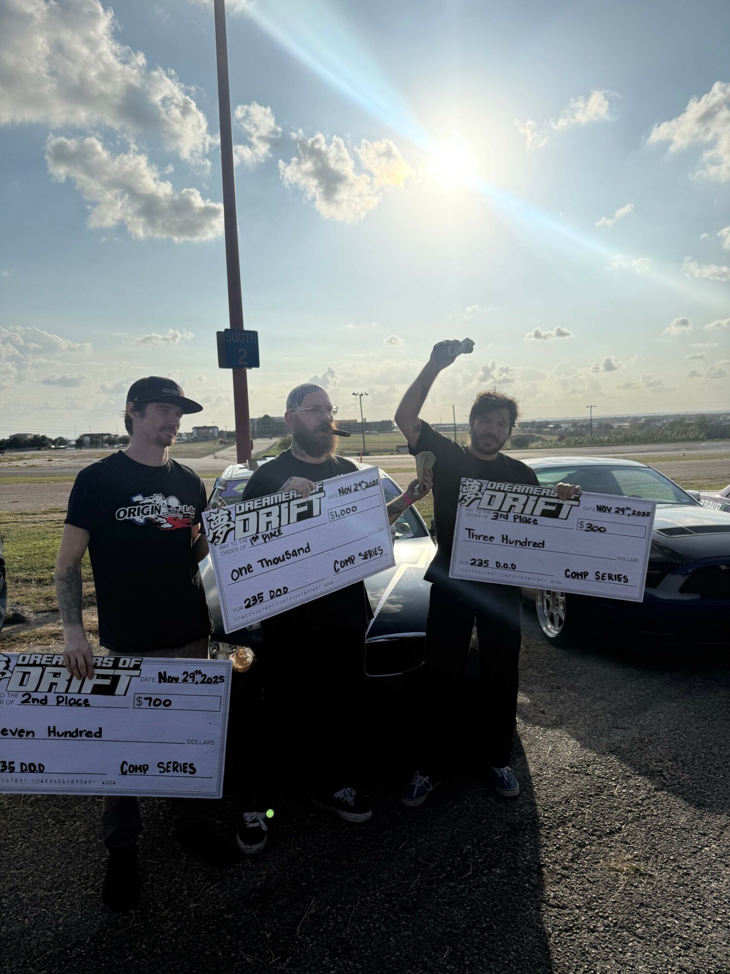 Three people holding large checks outdoors with a bright sun and partly cloudy sky, standing on a paved area.