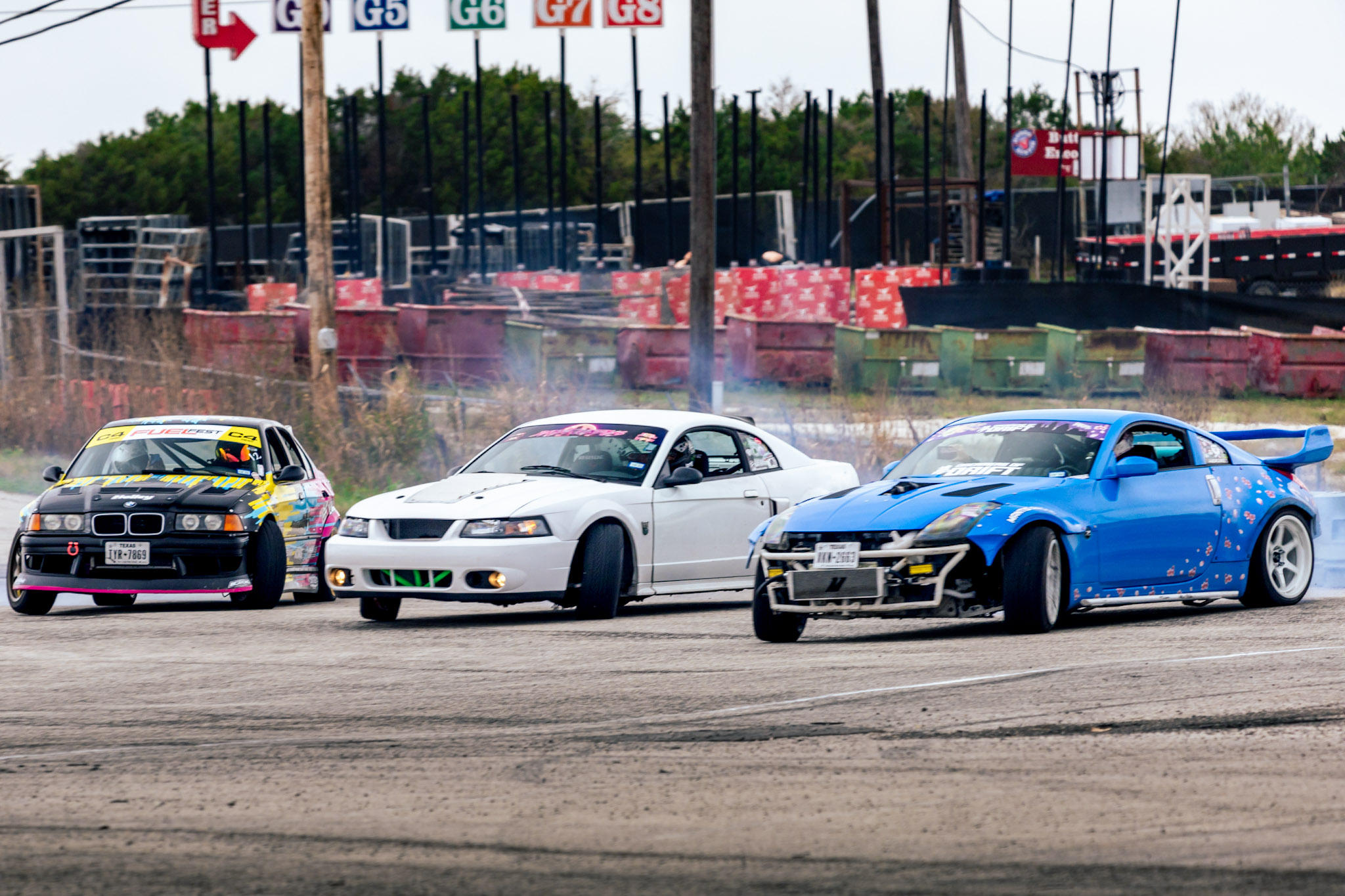 Three race cars on a track, with colorful barriers and flags in the background.