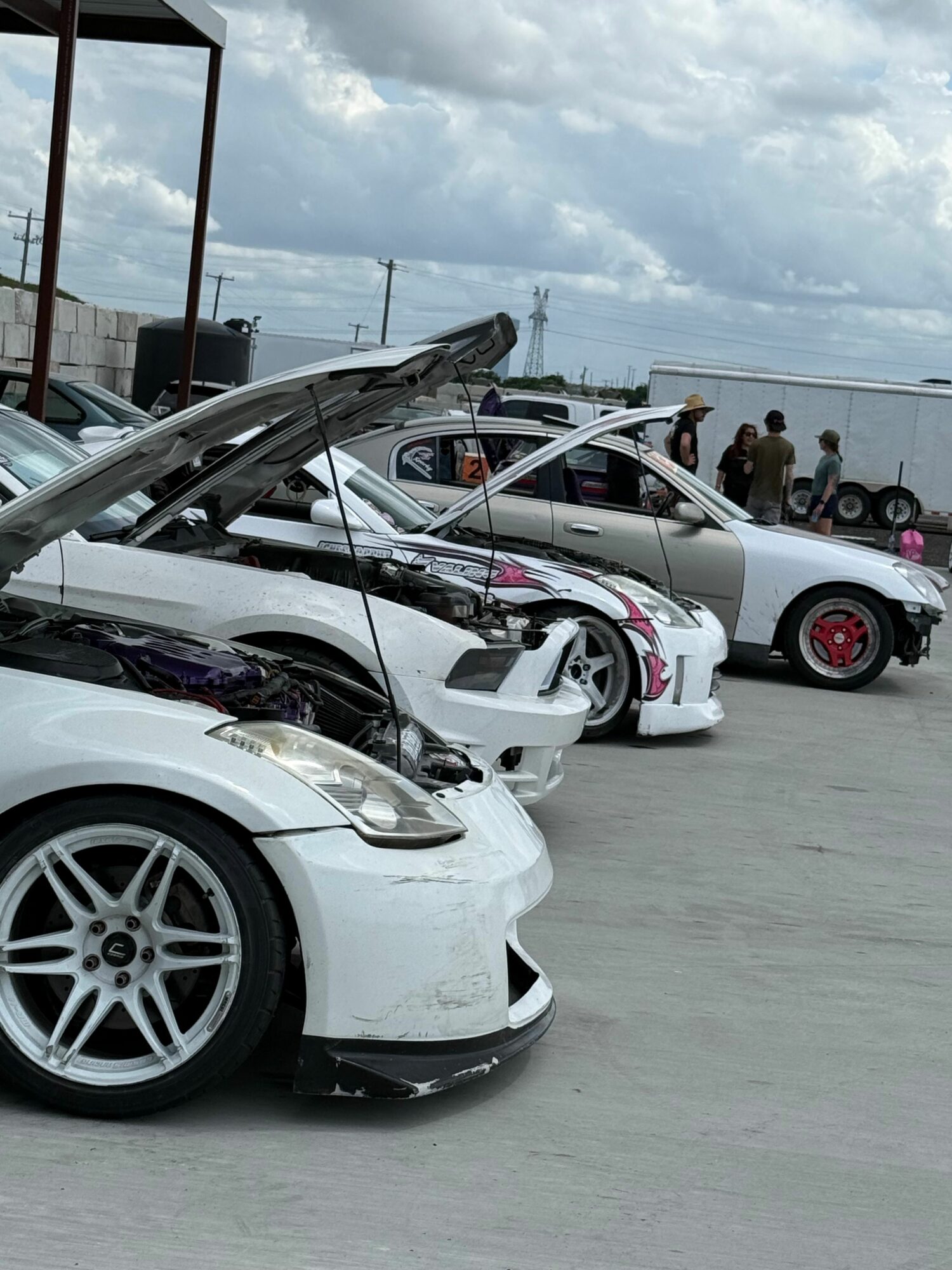 Multiple damaged white cars parked outdoors, some with open hoods and visible damage, under cloudy sky.