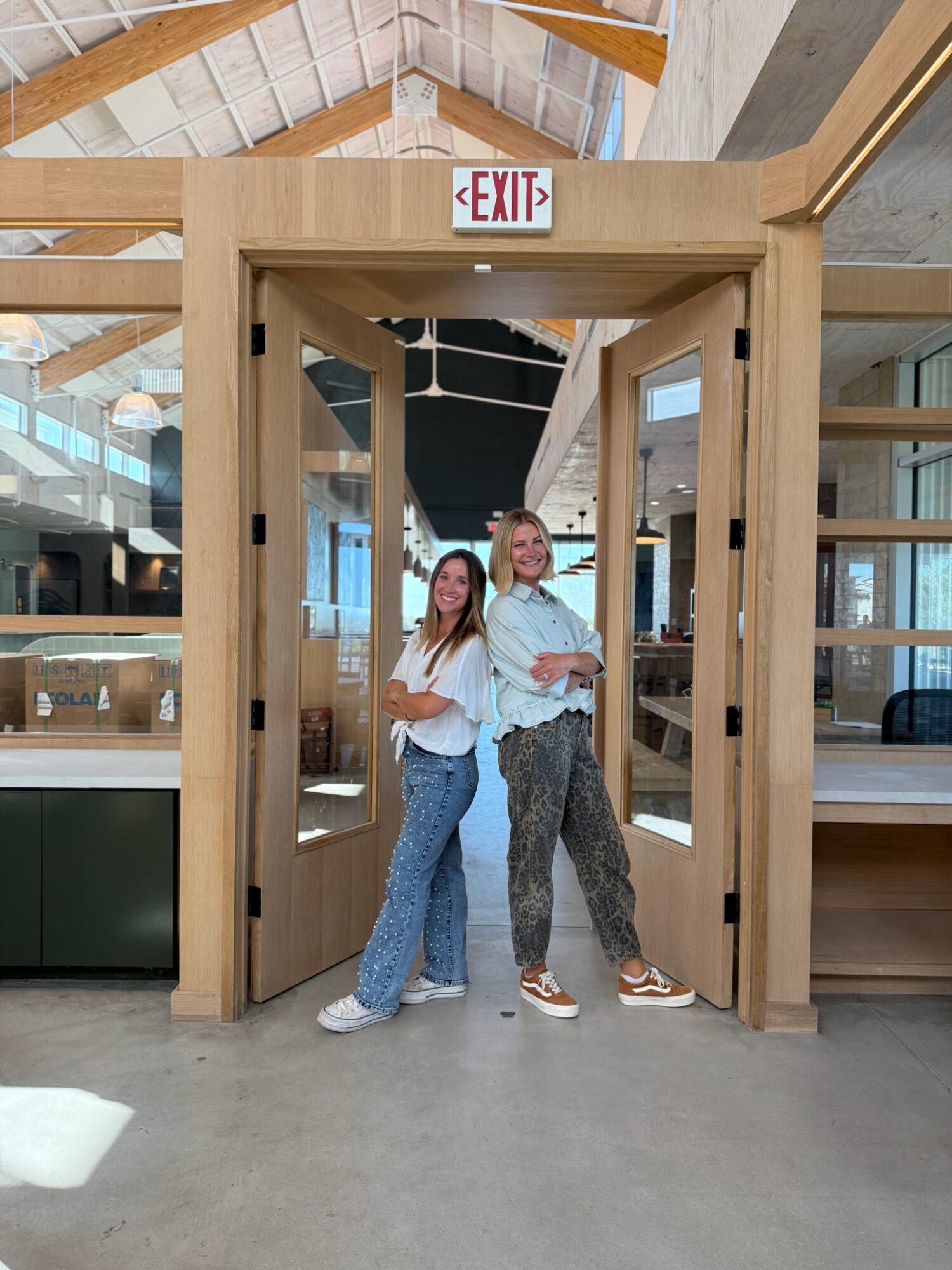 Two people stand in a doorway with an exit sign above, inside a modern building with wooden and glass elements.