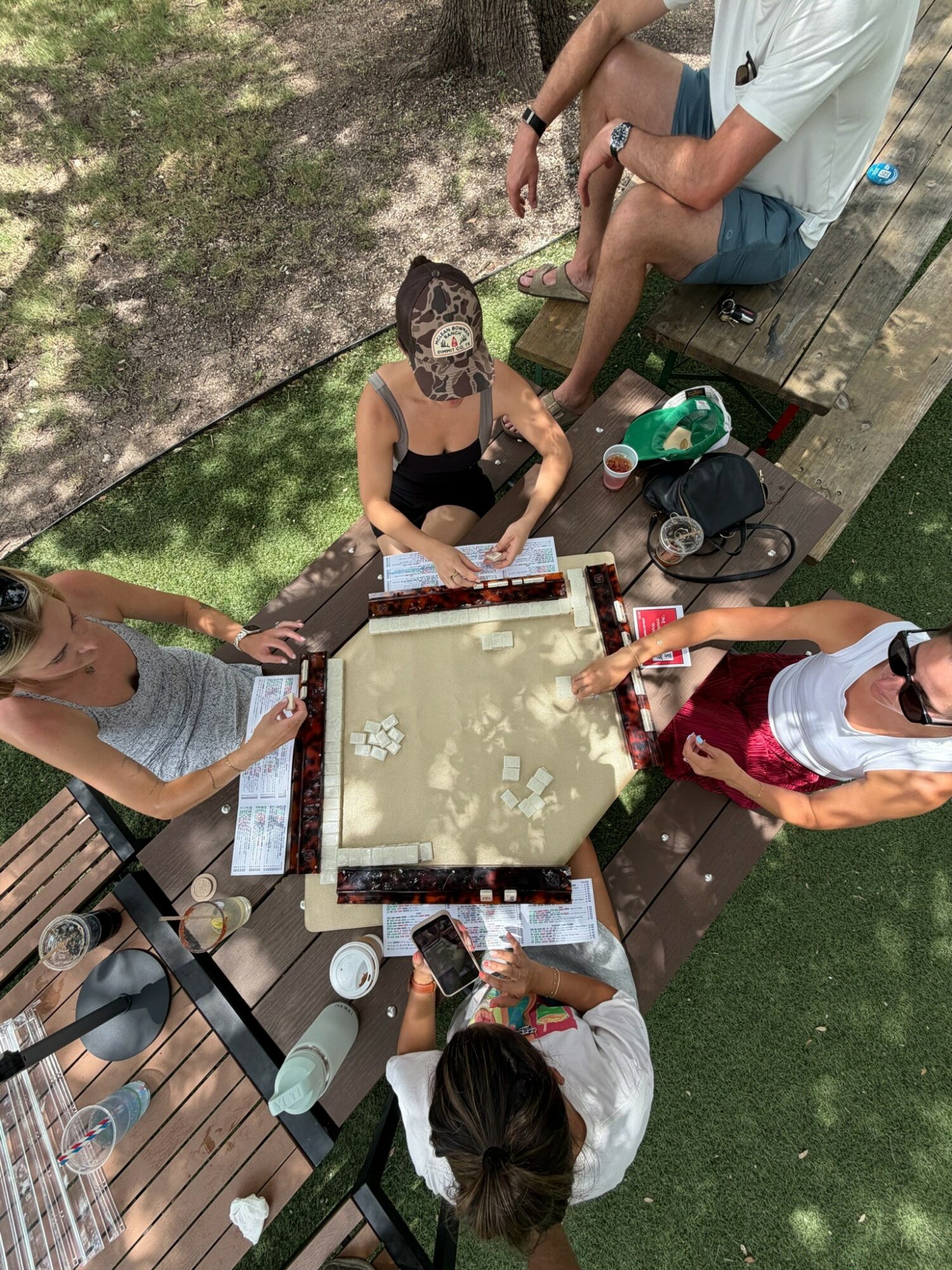 Six people sitting and standing around a table outdoors, playing a game, with drinks and snacks on the table.