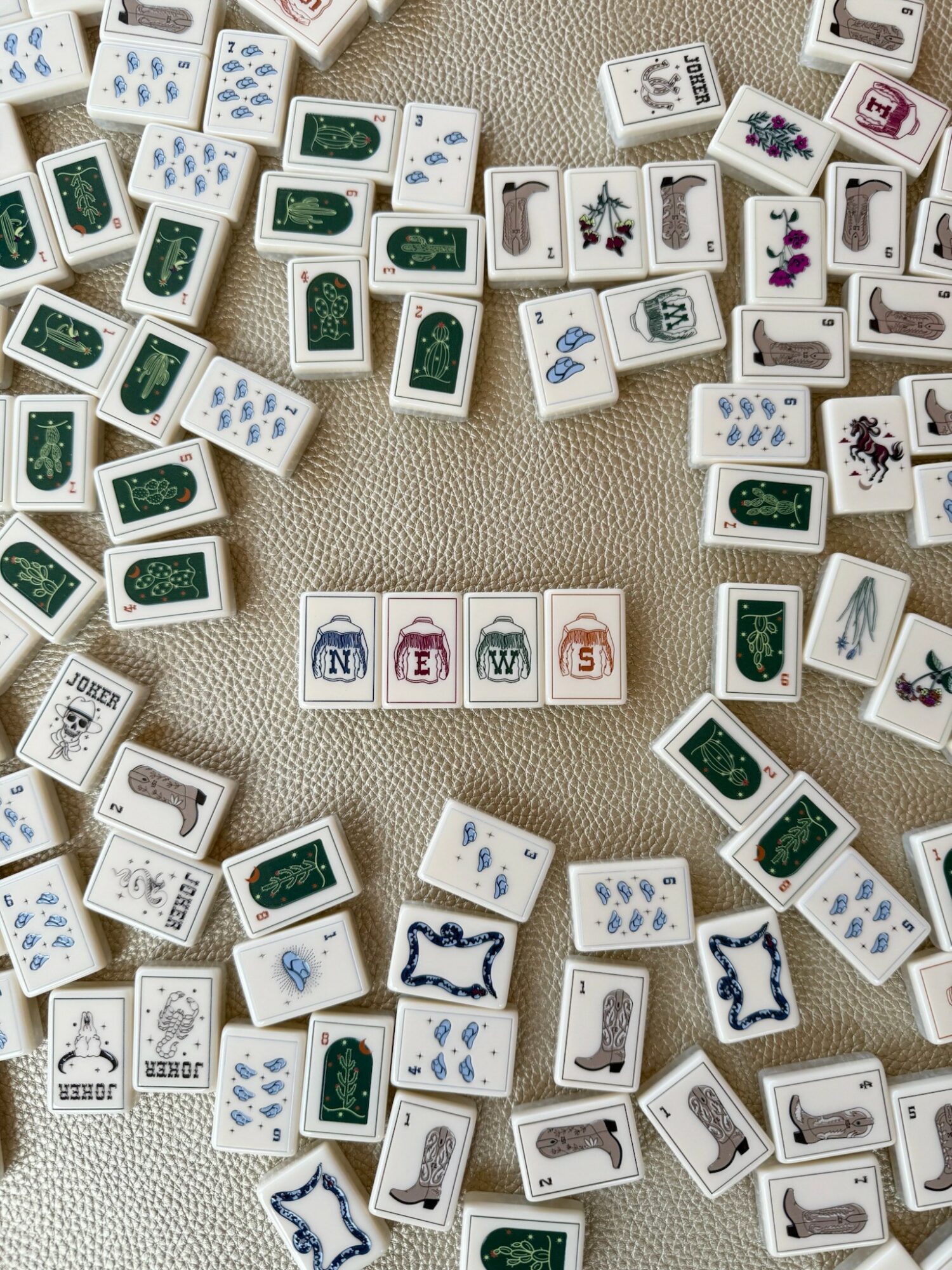Mahjong tiles scattered on a beige surface, some showing characters, bamboo, and symbols, with a row of letter tiles spelling 'NEWS' in the center.