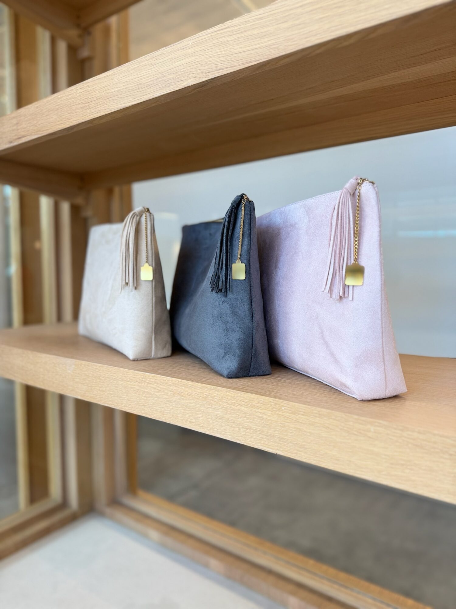 Three small fabric pouches in white, blue, and pink with tassels and gold accents on a wooden shelf.