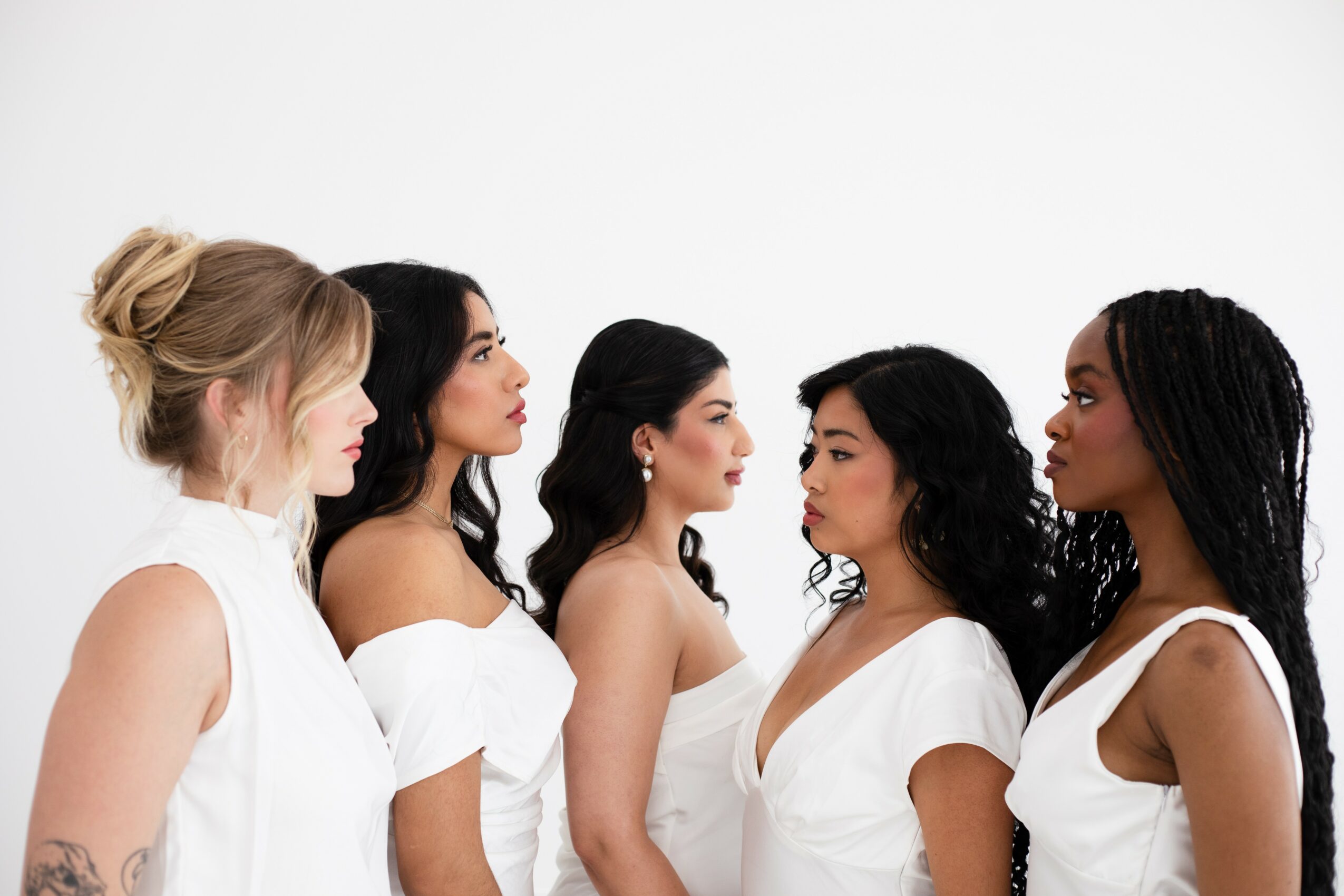 Five women standing in profile facing each other, all wearing white clothing, with a plain white background.