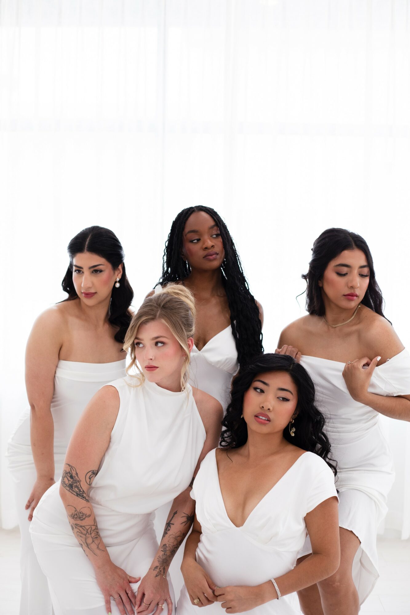 Five women in white dresses standing together against a bright background, looking in different directions.