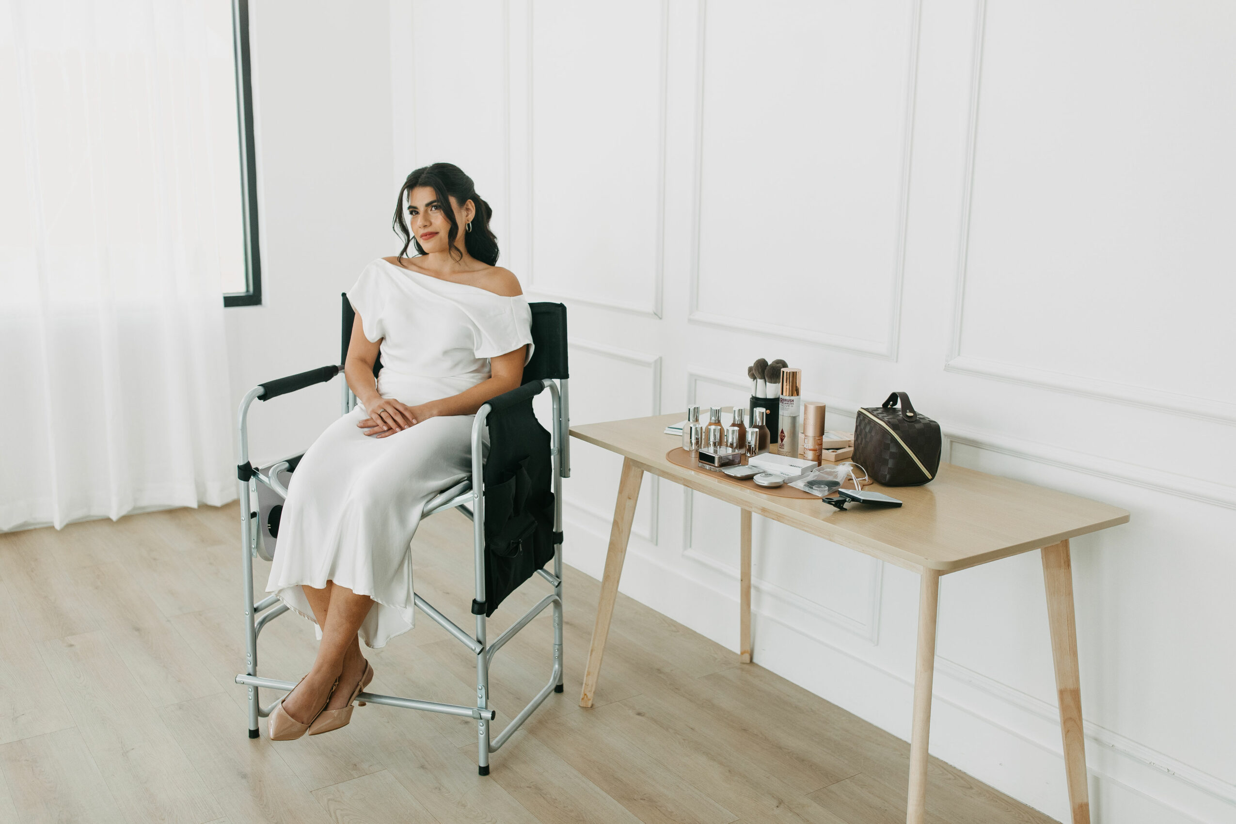 Woman in white dress sitting on a chair in a bright room with a table holding makeup items and a bag.