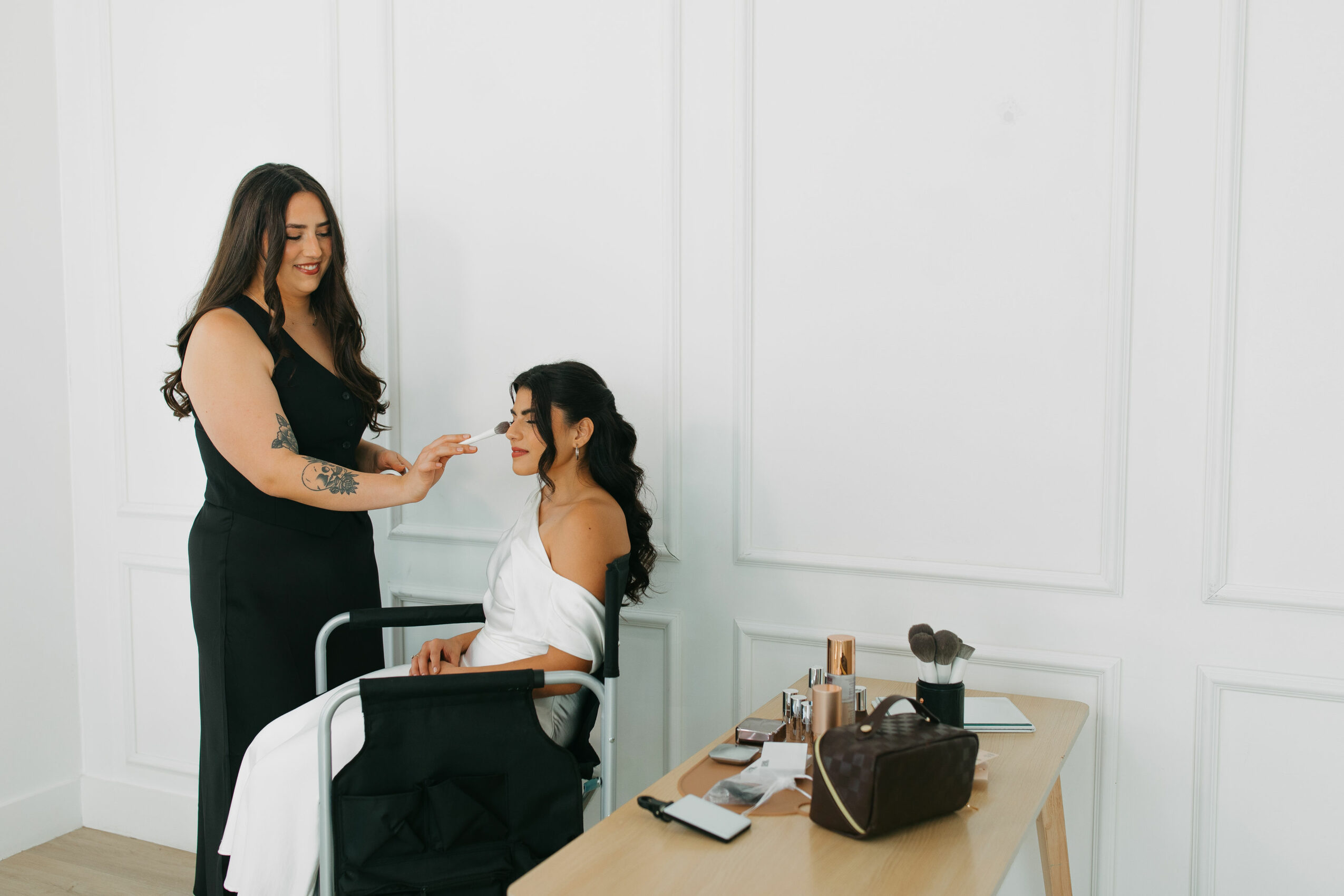 Two women in a room with white walls, one standing and the other sitting, engaging in conversation or a demonstration.