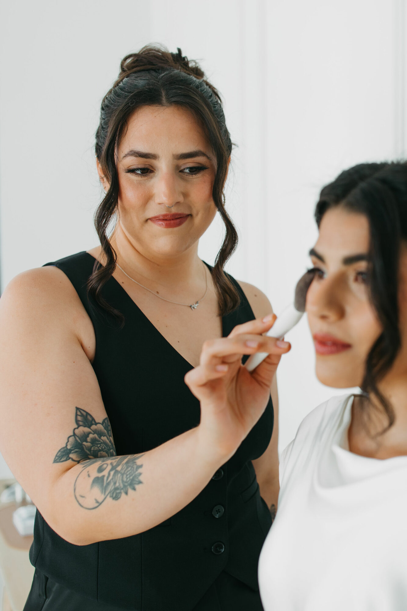A makeup artist applies makeup to a woman with dark hair, tattoos on her arm, and a black top.