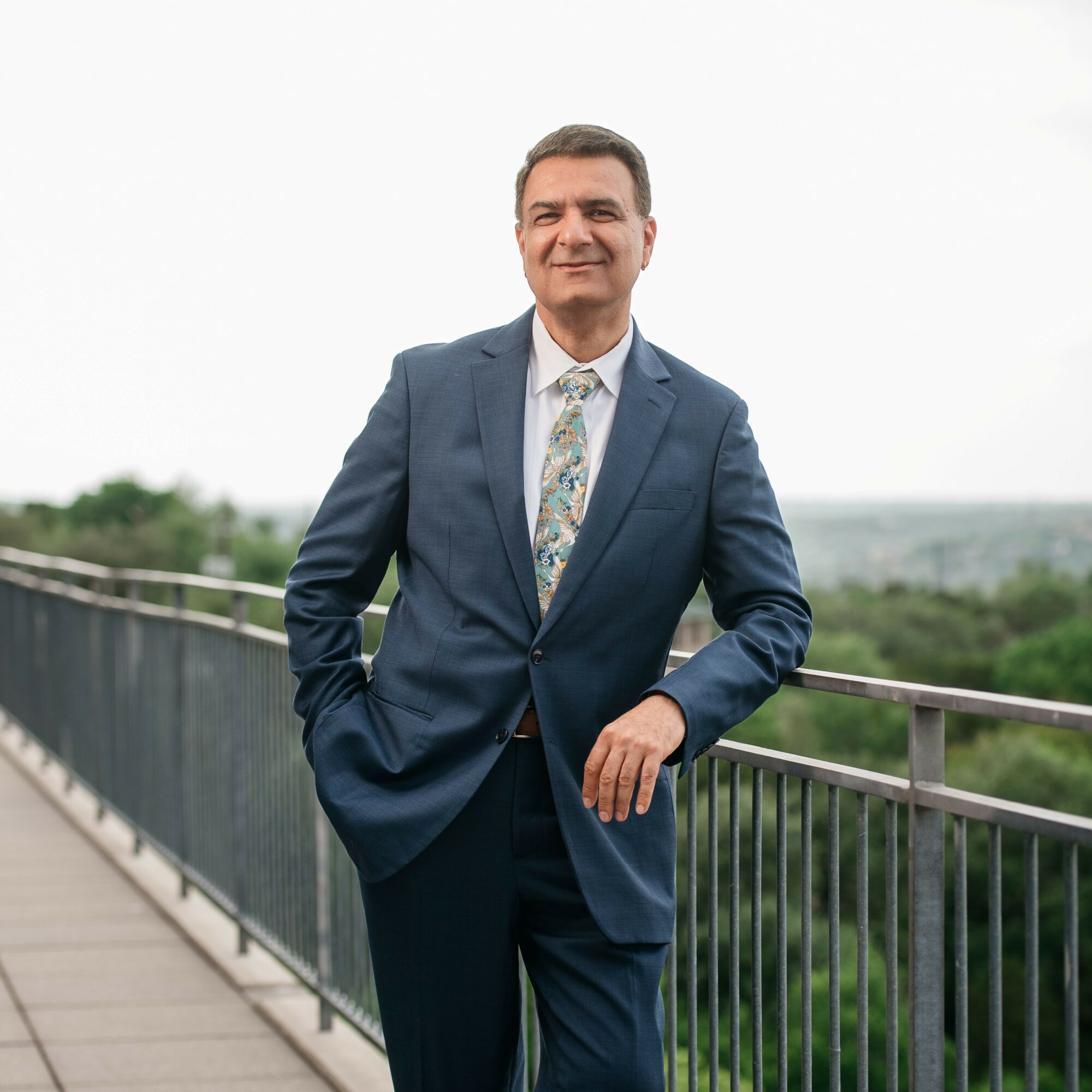 Man in a blue suit and patterned tie standing outdoors on a balcony with a railing, smiling, with greenery and sky in background.