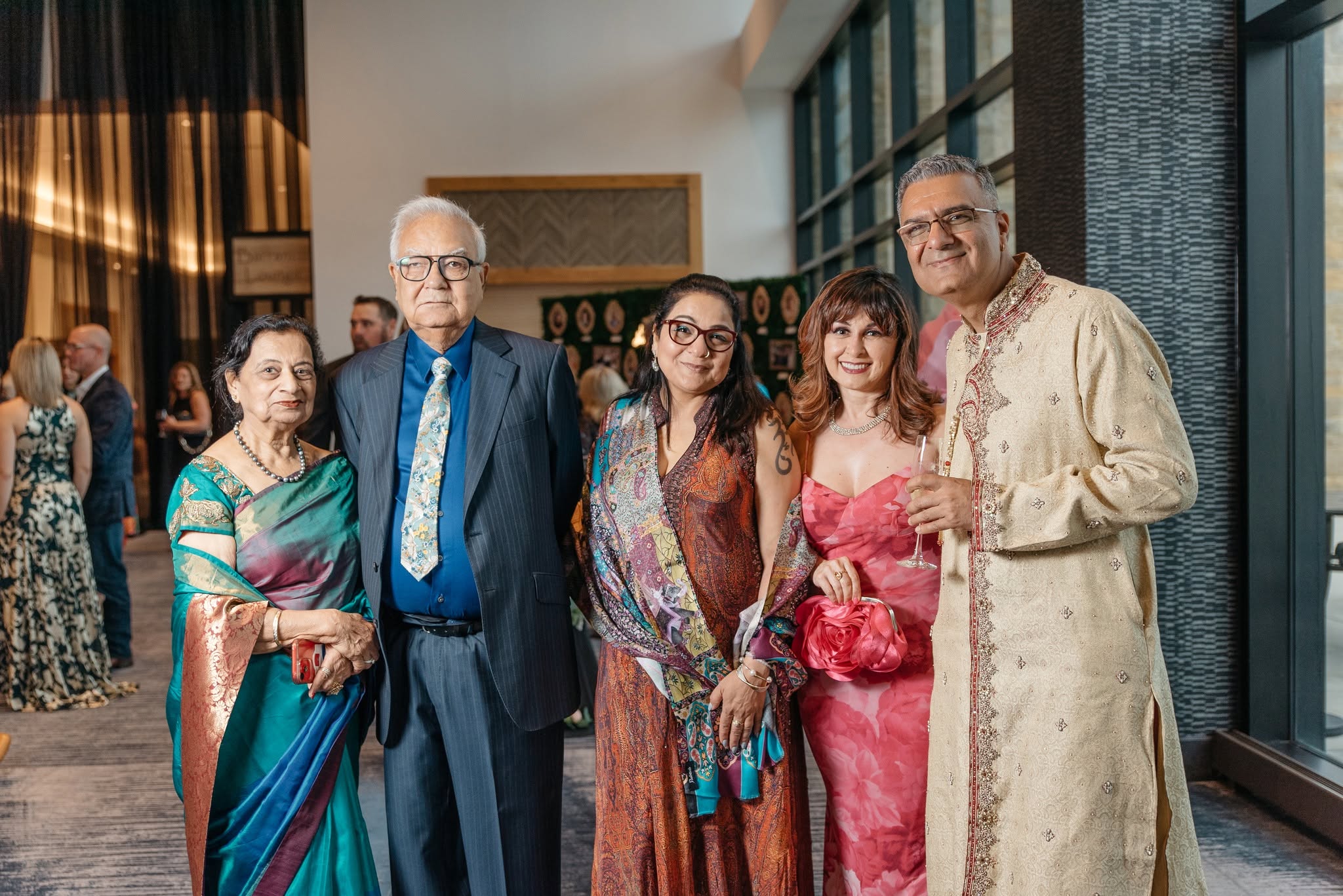 Group of five people smiling indoors, dressed in formal attire, standing near large windows with city view.