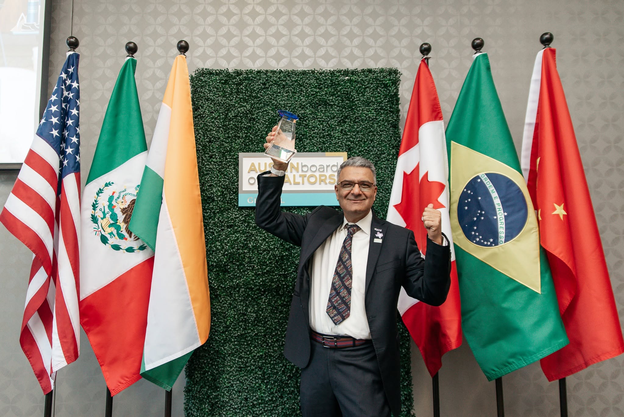 Man in suit smiling and holding a small flag, standing in front of flags from the US, Mexico, Ireland, Canada, Brazil, and China.