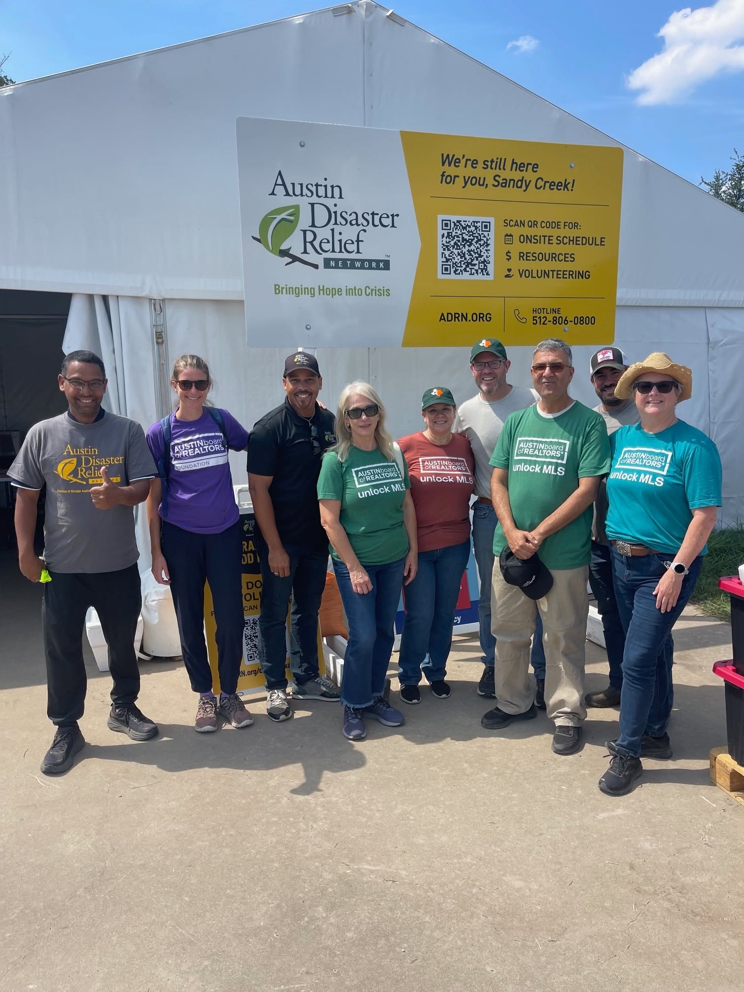 Group of eight people standing outside a large white tent with Austin Disaster Relief sign, sunny day.