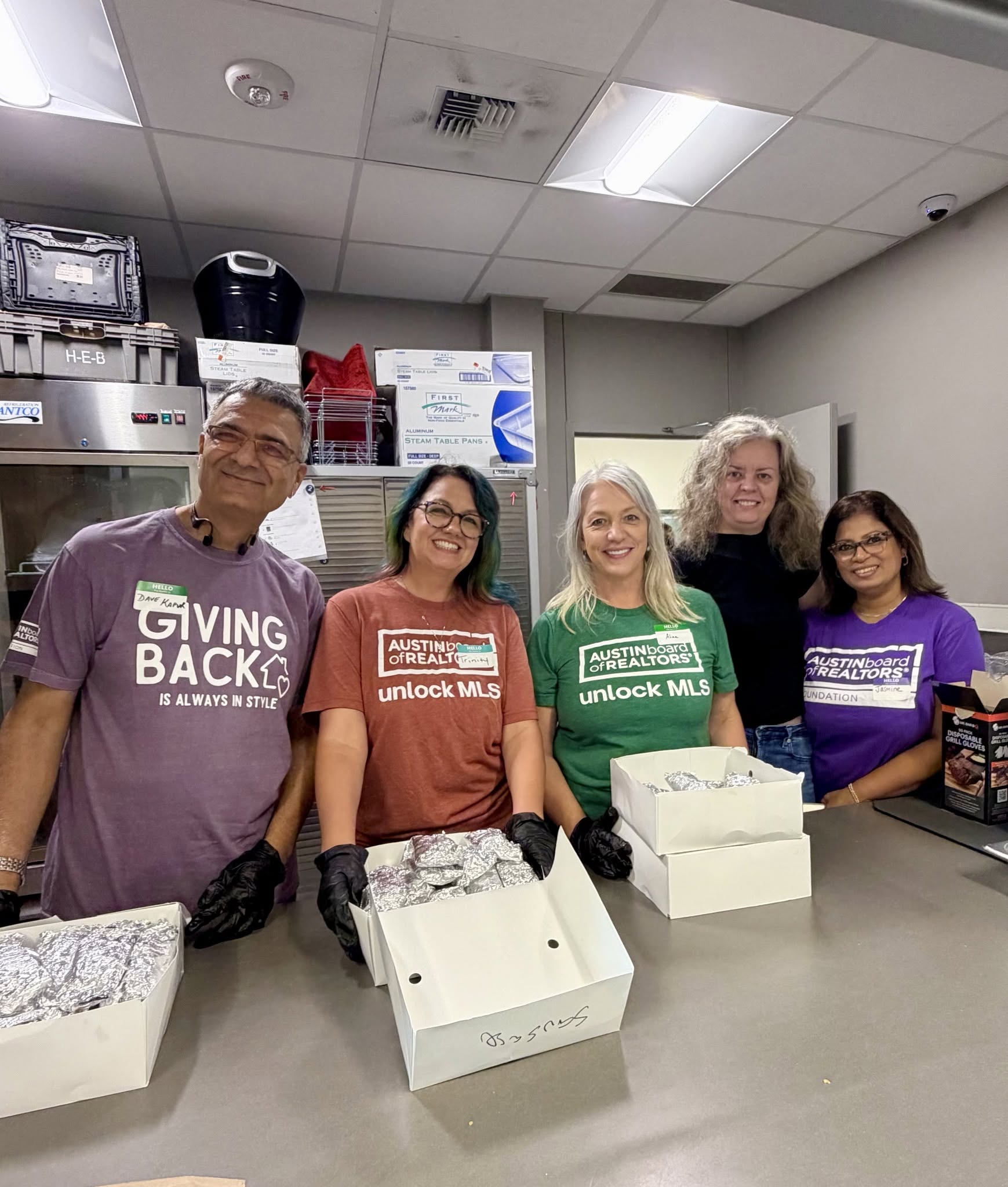 Five people standing behind a counter with boxes, smiling in a room with kitchen appliances and shelves.