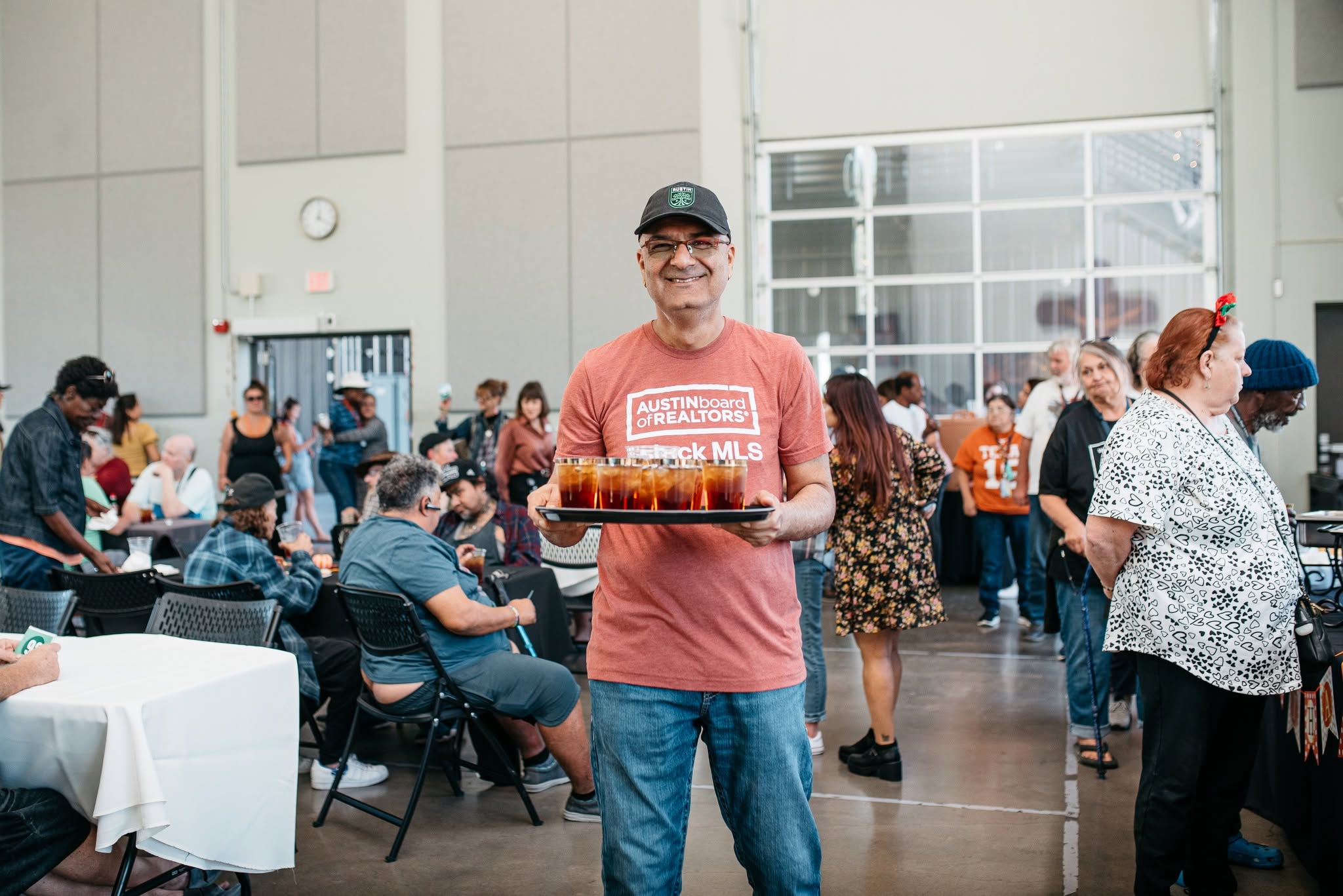 Man holding a tray with food at a crowded indoor event, smiling, wearing glasses, a cap, and casual clothing.