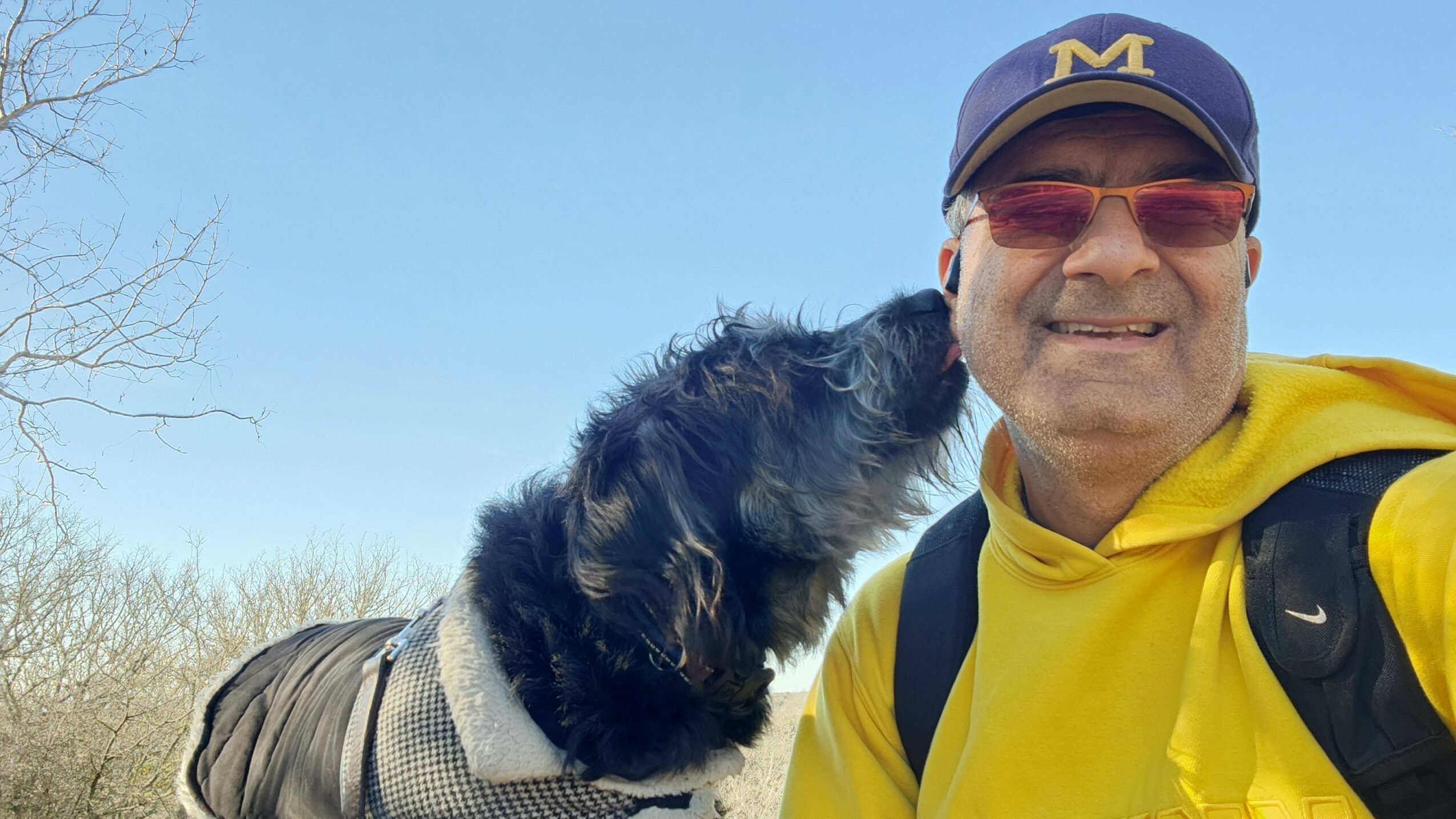Person wearing a yellow jacket and cap smiling outdoors with a dog licking their face.