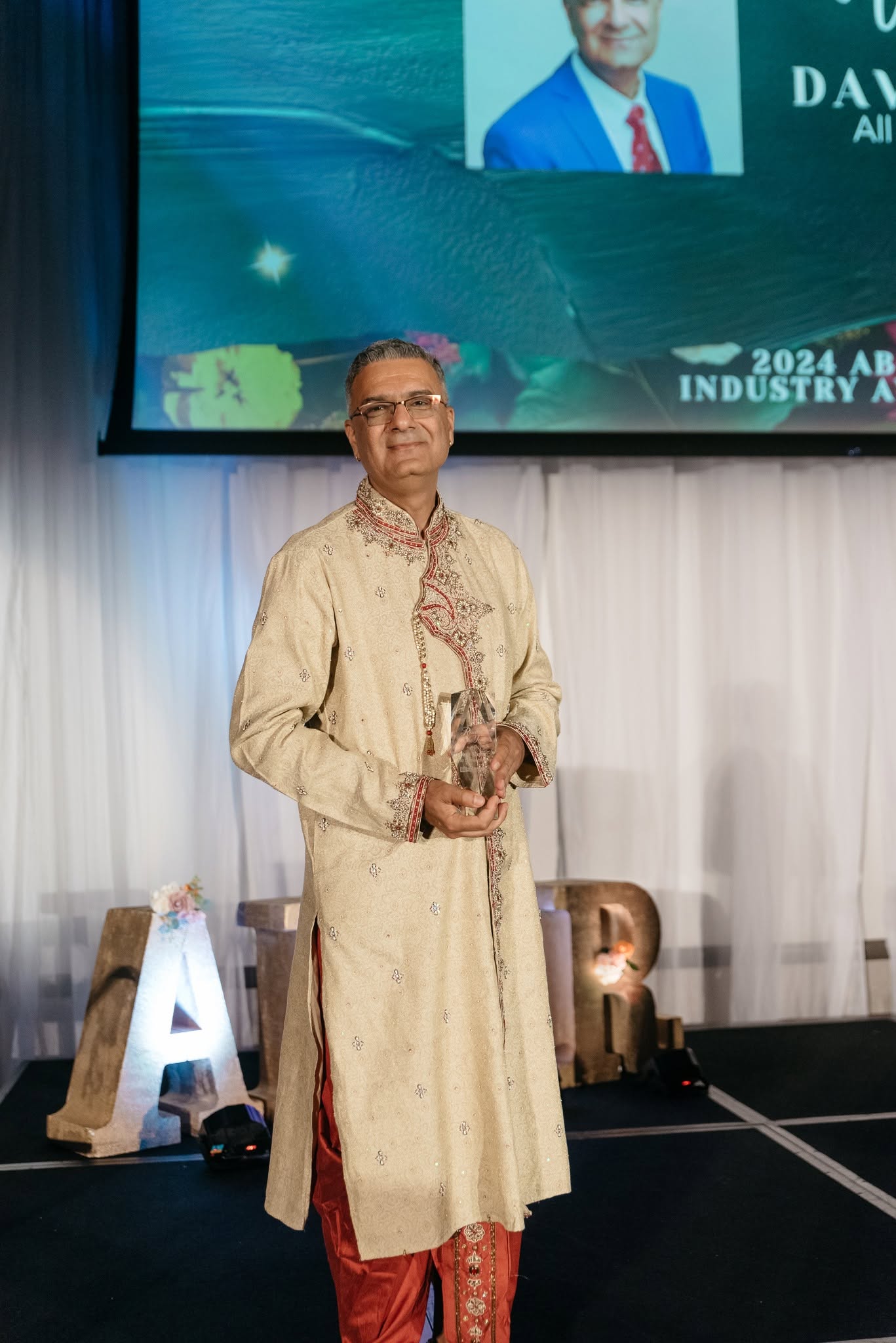 Man in traditional attire standing on stage holding an award, with a large screen and decorative letters behind him.