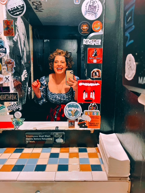 Woman with curly hair smiling in a mirror surrounded by stickers, taking a selfie in a decorated space.