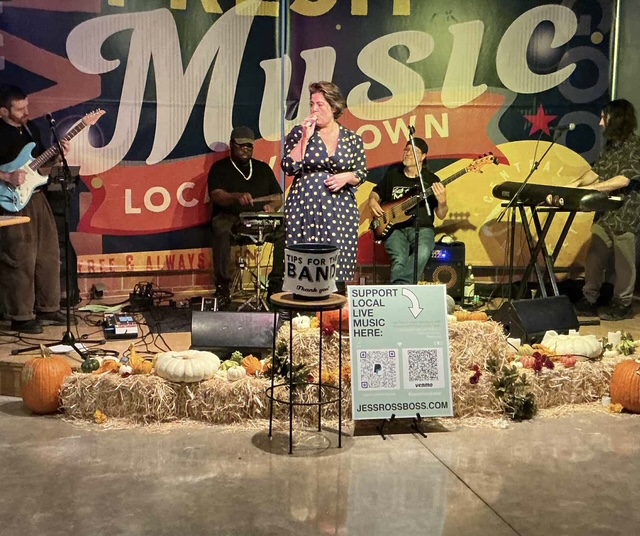 Group of musicians performing on stage with pumpkins and hay bales, woman singing into microphone, colorful backdrop, support sign.