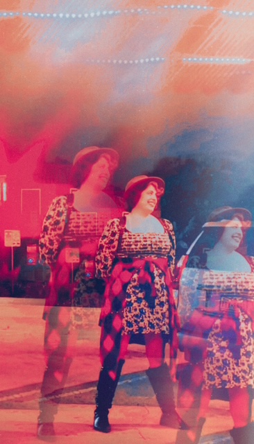 Three women in patterned dresses and hats perform on stage with colorful lighting and sky background.