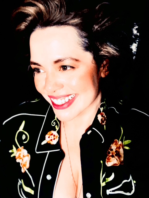 Smiling woman with dark hair, wearing floral embroidered black jacket, against a dark background.