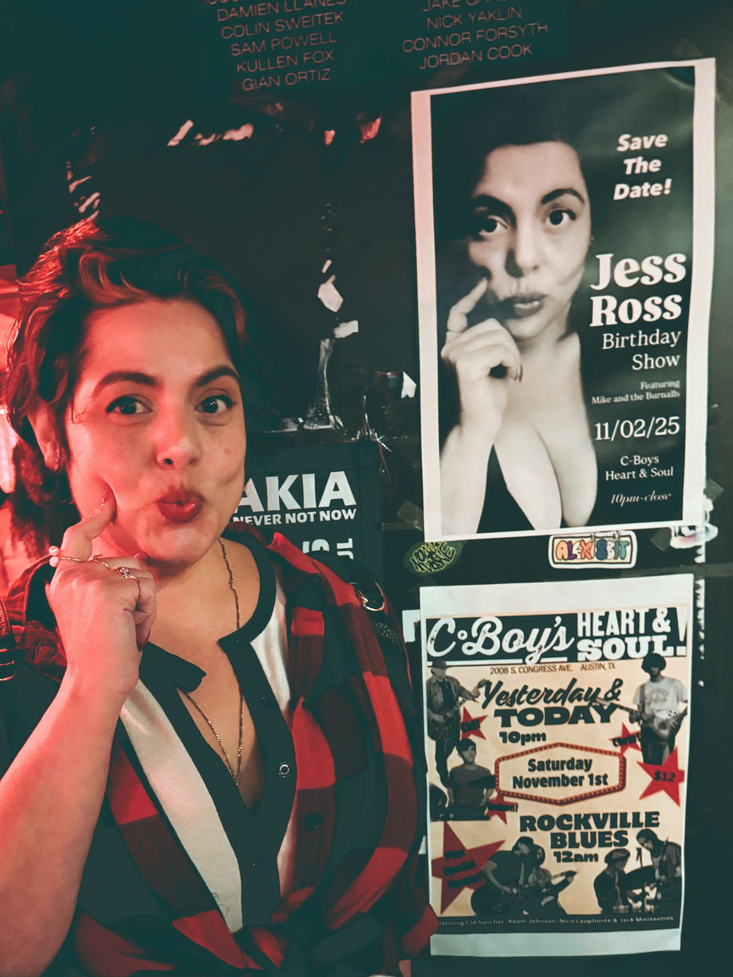 Young woman with red hair and plaid shirt making a playful face, standing next to a poster of Jess Ross's birthday show.