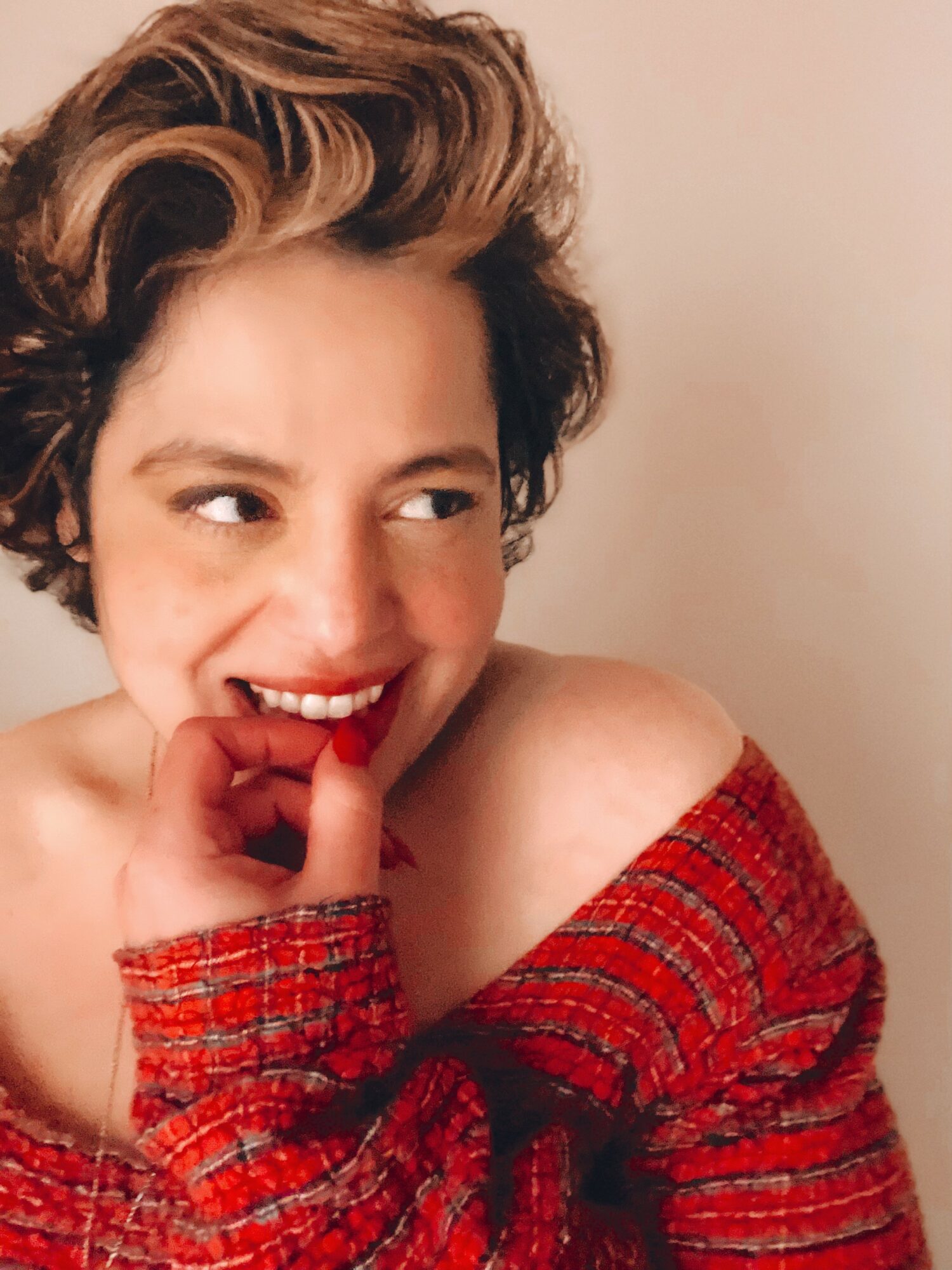 Woman with short, wavy hair smiling and biting her finger, wearing a red off-shoulder sweater.