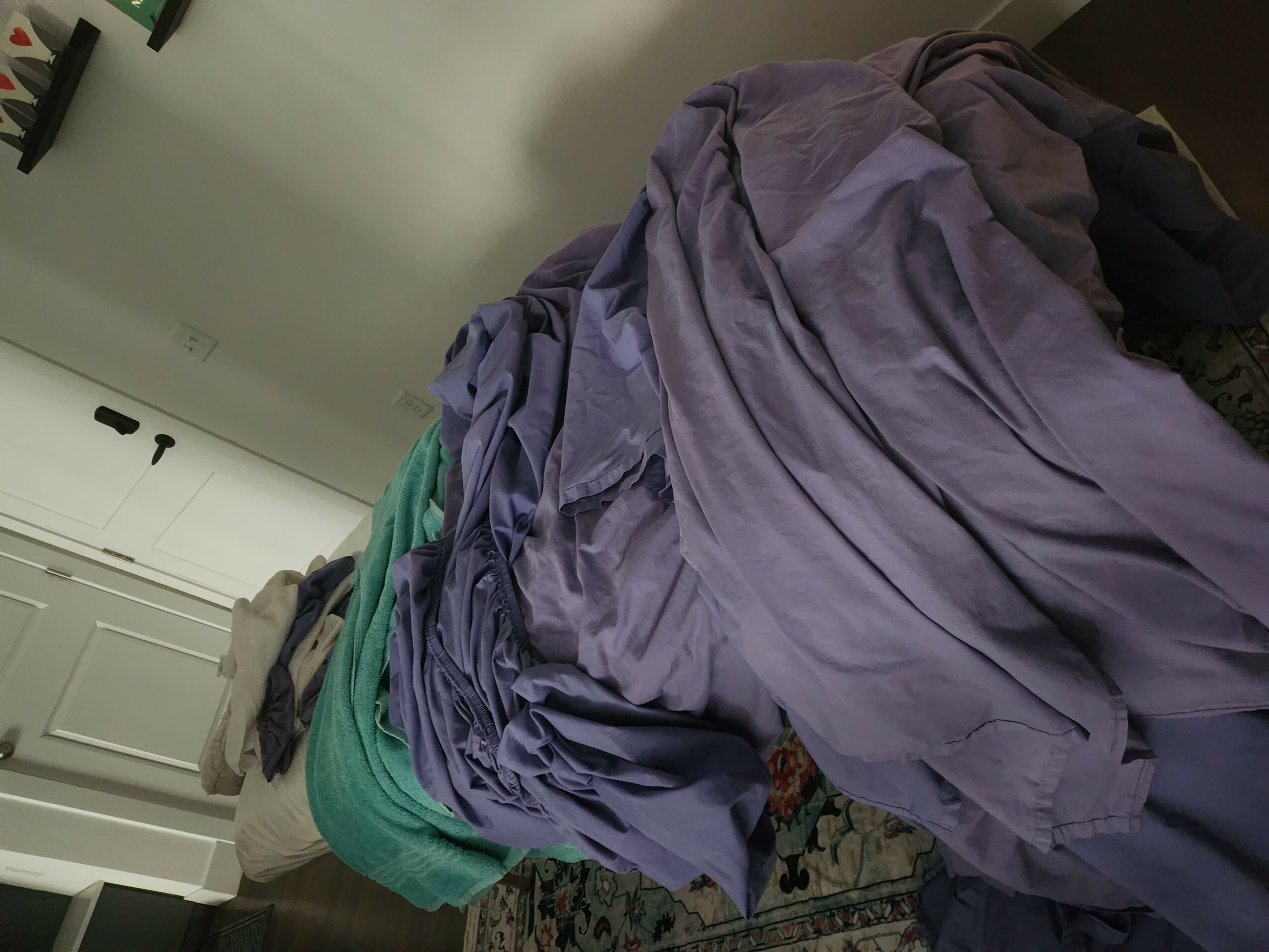 Clothes piled on a bed with a patterned blanket, a white door, and a light-colored wall in the background.