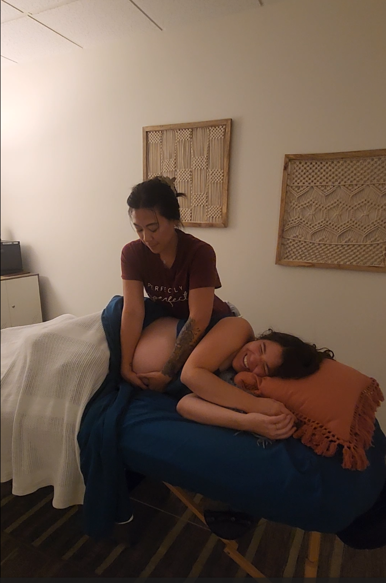 Person lying on bed with head on pillow, another person sitting on bed, adjusting their clothing, in a room with wall art.