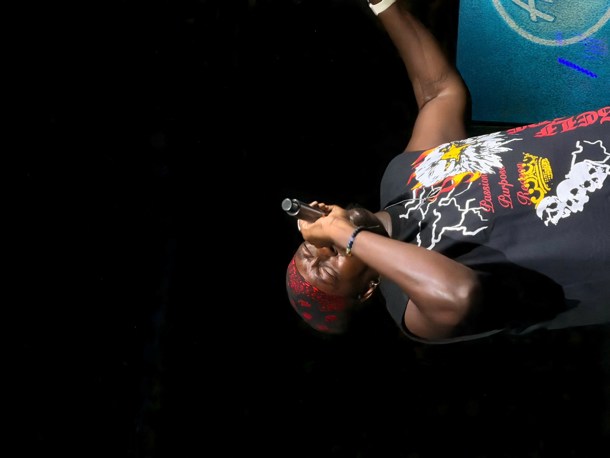 Person lying down, holding a gun, wearing a black shirt with colorful designs, against a dark background.