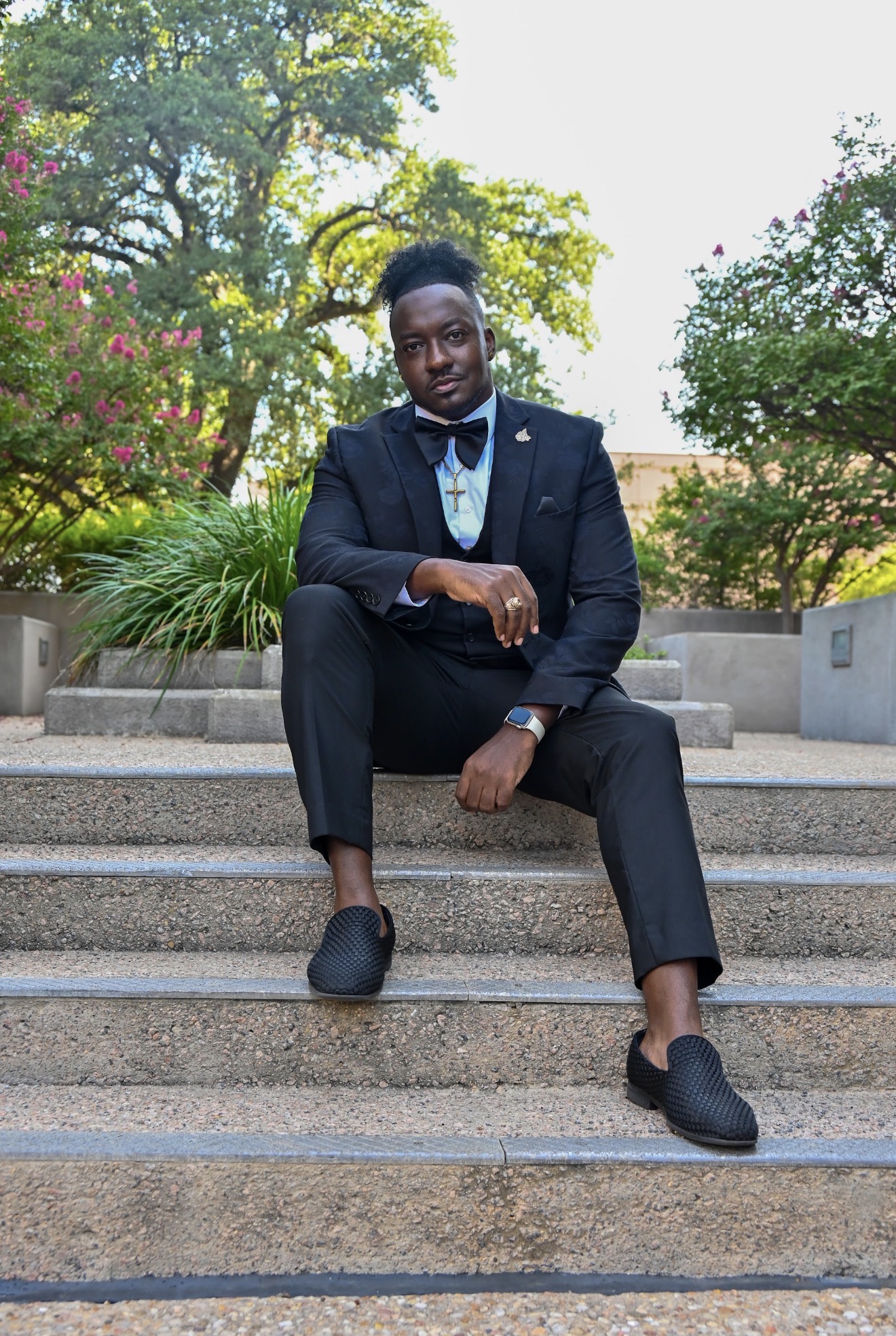 Person in formal suit sitting on outdoor steps with trees and plants in background.